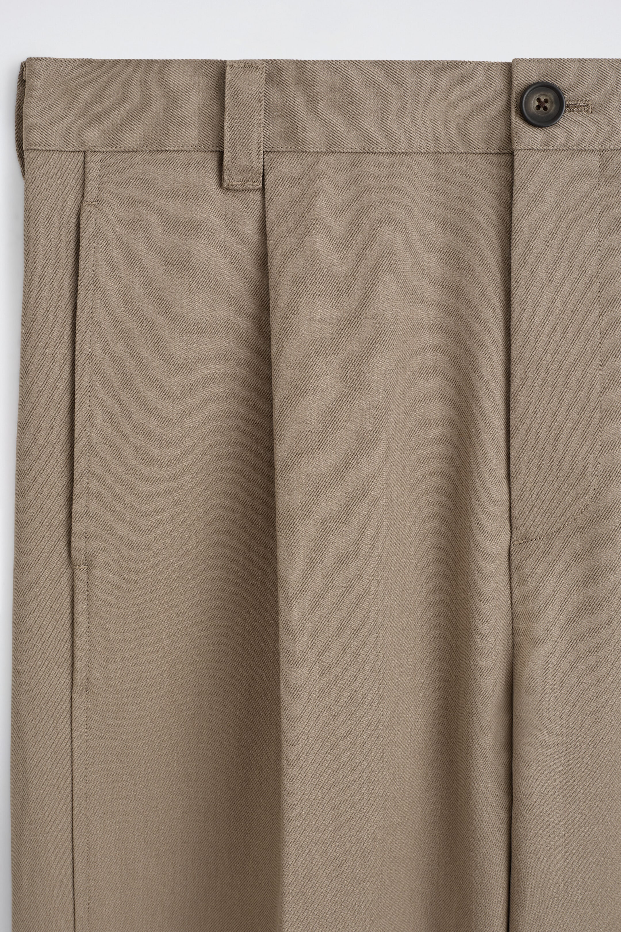 Estos pantalones color topo se presentan en una vista frontal de primer plano de la zona de la cintura, mostrando el tejido de sarga, un único pliegue delantero, un bolsillo lateral vertical, trabillas para el cinturón y un cierre de botón oscuro. Estos pantalones color topo se presentan en una vista frontal de primer plano de la zona de la cintura, mostrando el tejido de sarga, un único pliegue delantero, un bolsillo lateral vertical, trabillas para el cinturón y un cierre de botón oscuro.