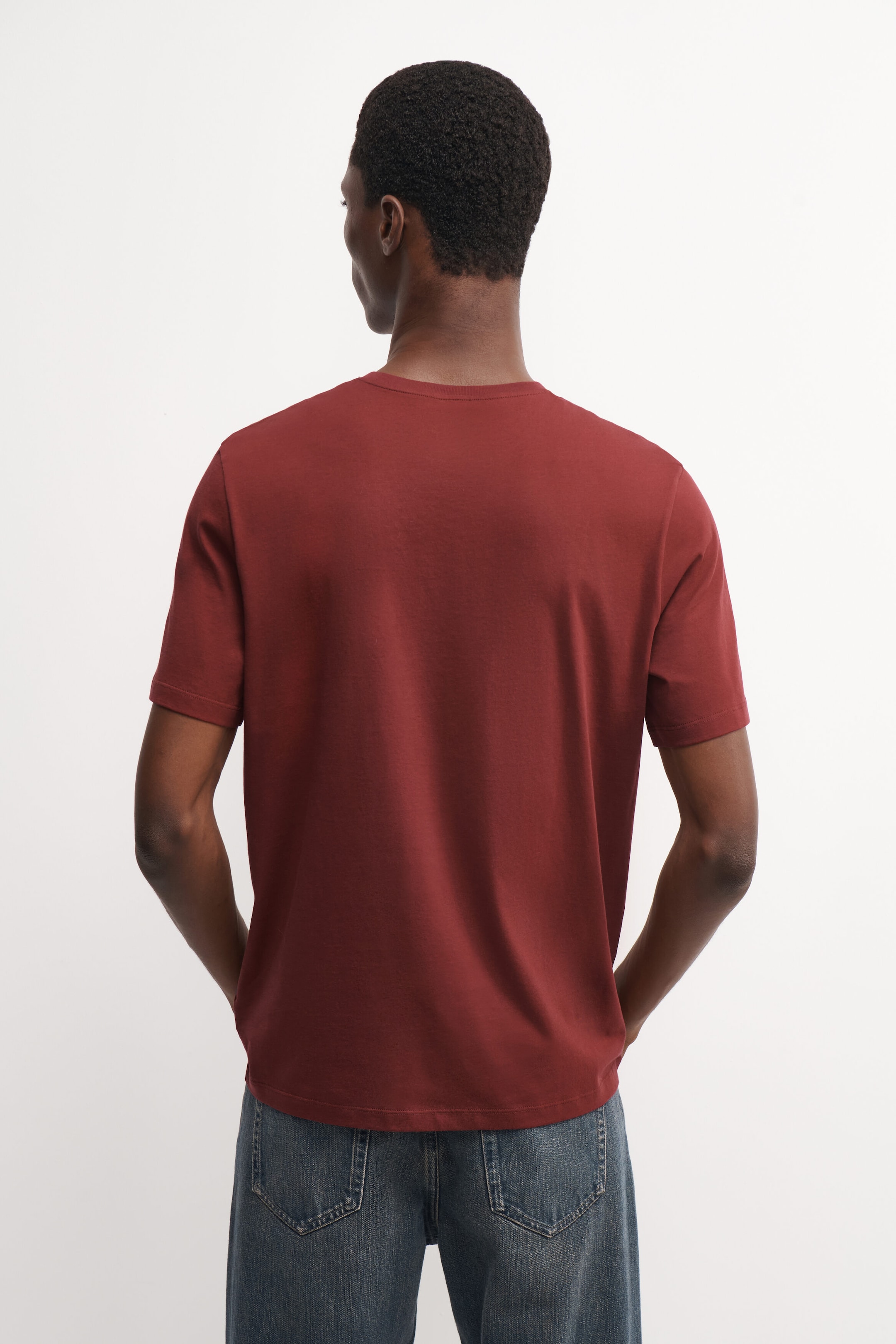 The model is wearing a dark red cotton crew-neck T-shirt, standing with their back to the camera. The short-sleeved, regular-fit T-shirt is styled with blue jeans. The model is wearing a dark red cotton crew-neck T-shirt, standing with their back to the camera. The short-sleeved, regular-fit T-shirt is styled with blue jeans.