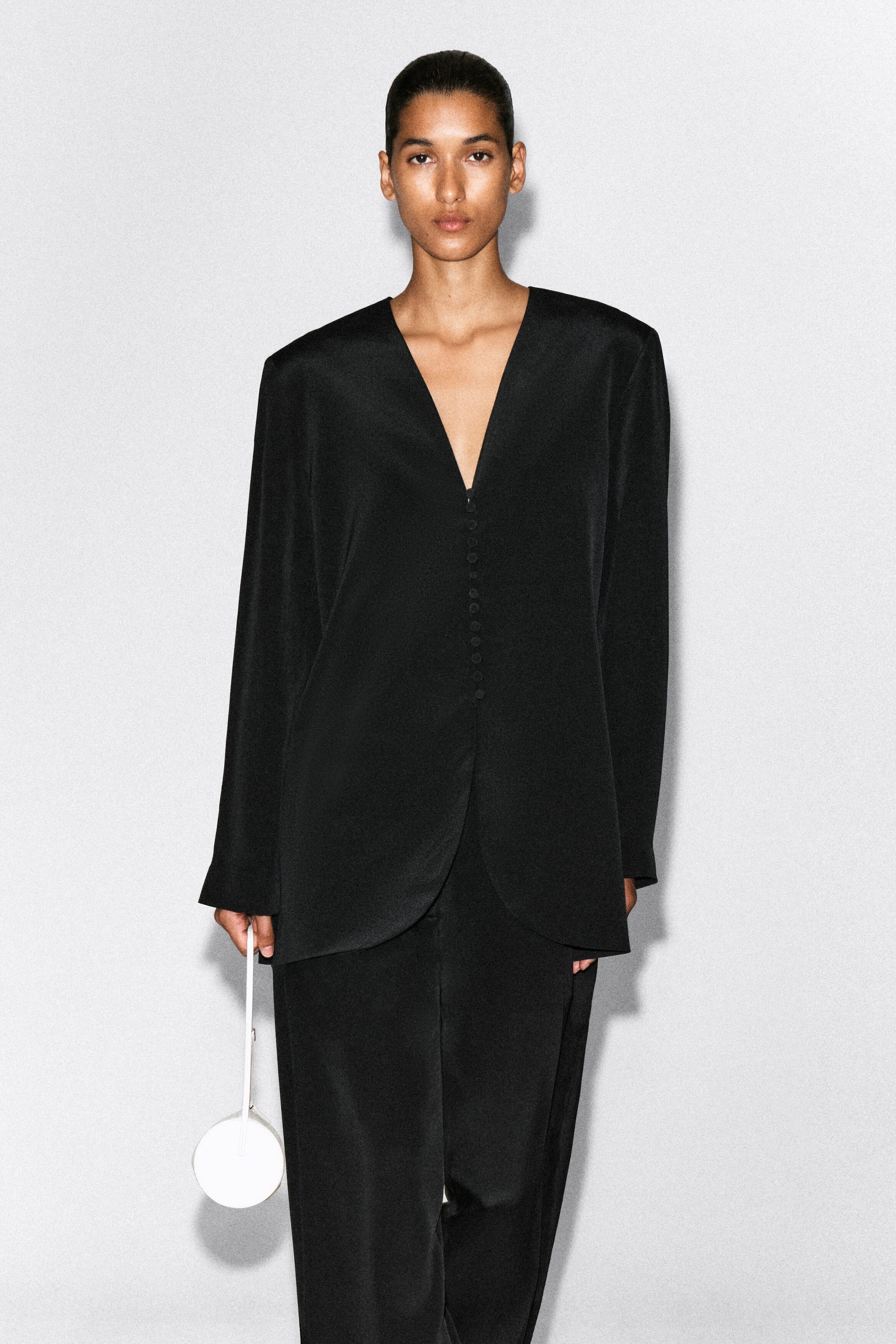 Von vorn präsentiert trägt ein Model einen kragenlosen, schmal geschnittenen schwarzen Seidenblazer mit tiefem V-Ausschnitt und einem mittigen Verschluss mit kleinen, stoffbezogenen Knöpfen. Der Blazer wird mit einer passenden schwarzen Hose kombiniert, und das Model hält eine kleine, runde weiße Tasche. Von vorn präsentiert trägt ein Model einen kragenlosen, schmal geschnittenen schwarzen Seidenblazer mit tiefem V-Ausschnitt und einem mittigen Verschluss mit kleinen, stoffbezogenen Knöpfen. Der Blazer wird mit einer passenden schwarzen Hose kombiniert, und das Model hält eine kleine, runde weiße Tasche.