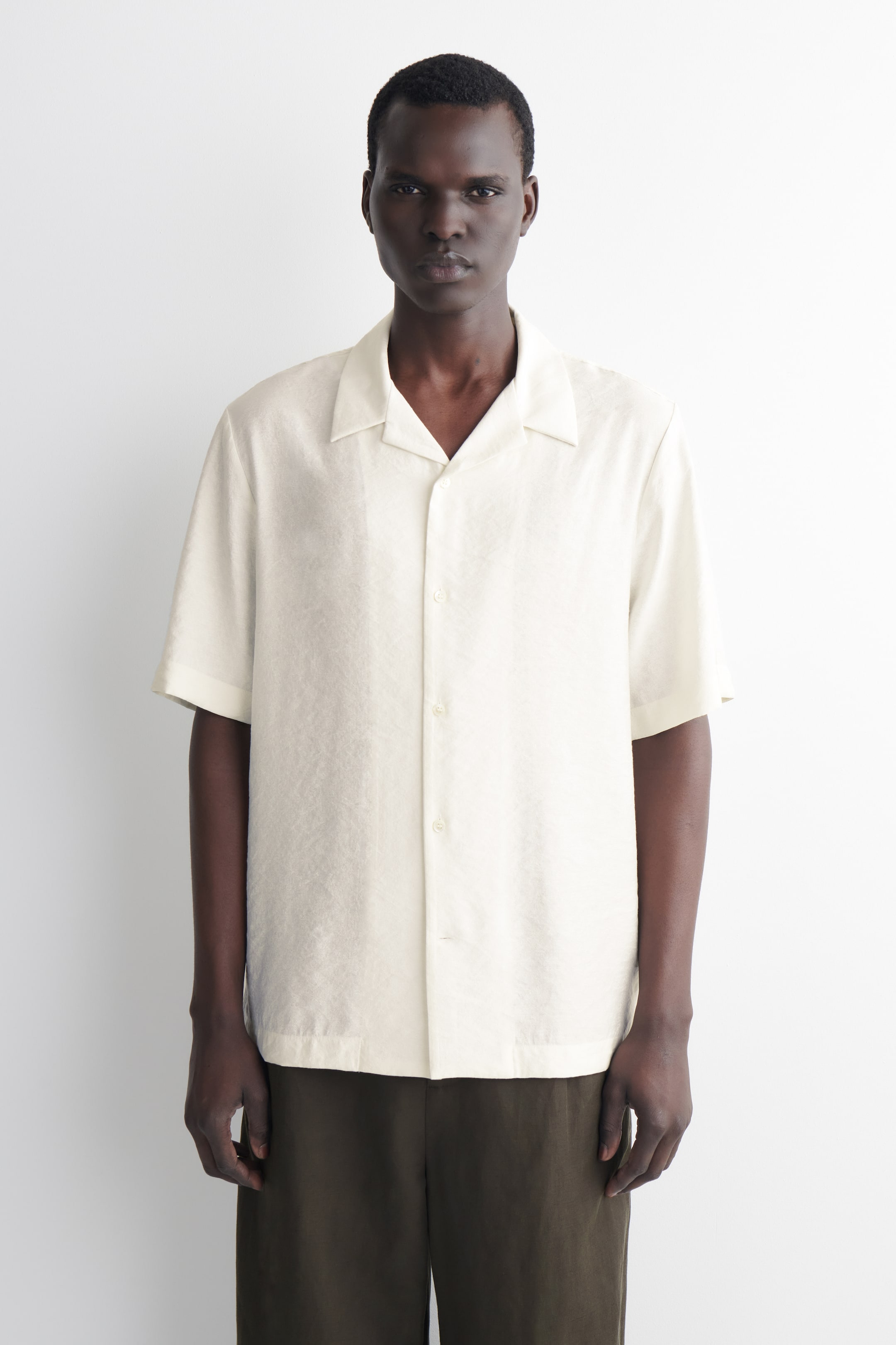 Ver imagen más grande: La camisa de manga corta en color crema se muestra de frente, con un cuello tipo campamento, cierre de botones en la parte delantera y un sutil patrón texturizado a lo largo de su tejido.