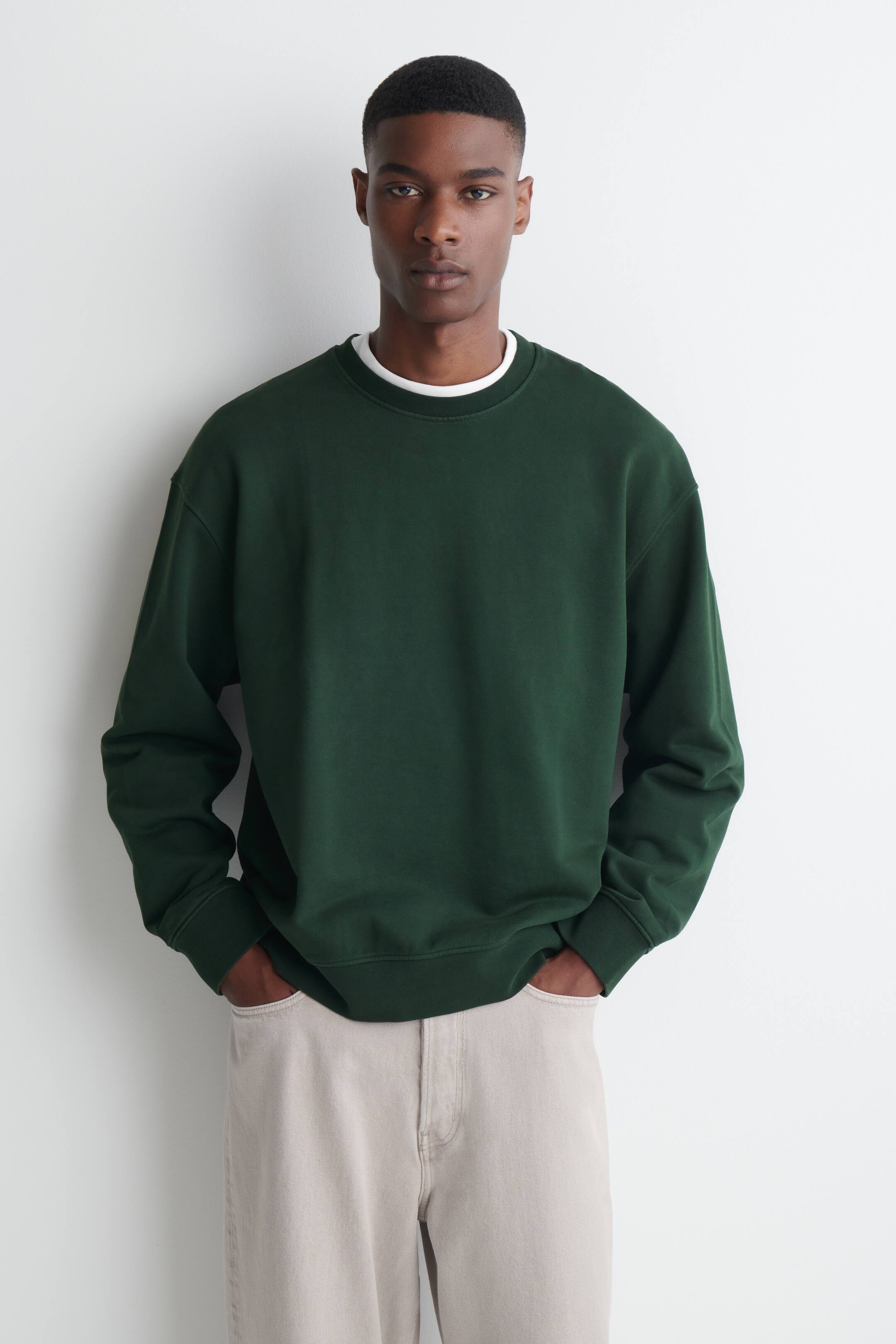 Agrandir l'image: Un sweat-shirt col rond vert foncé en coton délavé est porté par le modèle par-dessus un t-shirt blanc superposé. Le sweat-shirt présente des finitions côtelées, et le modèle se tient debout, les mains glissées dans les poches d’un pantalon clair.