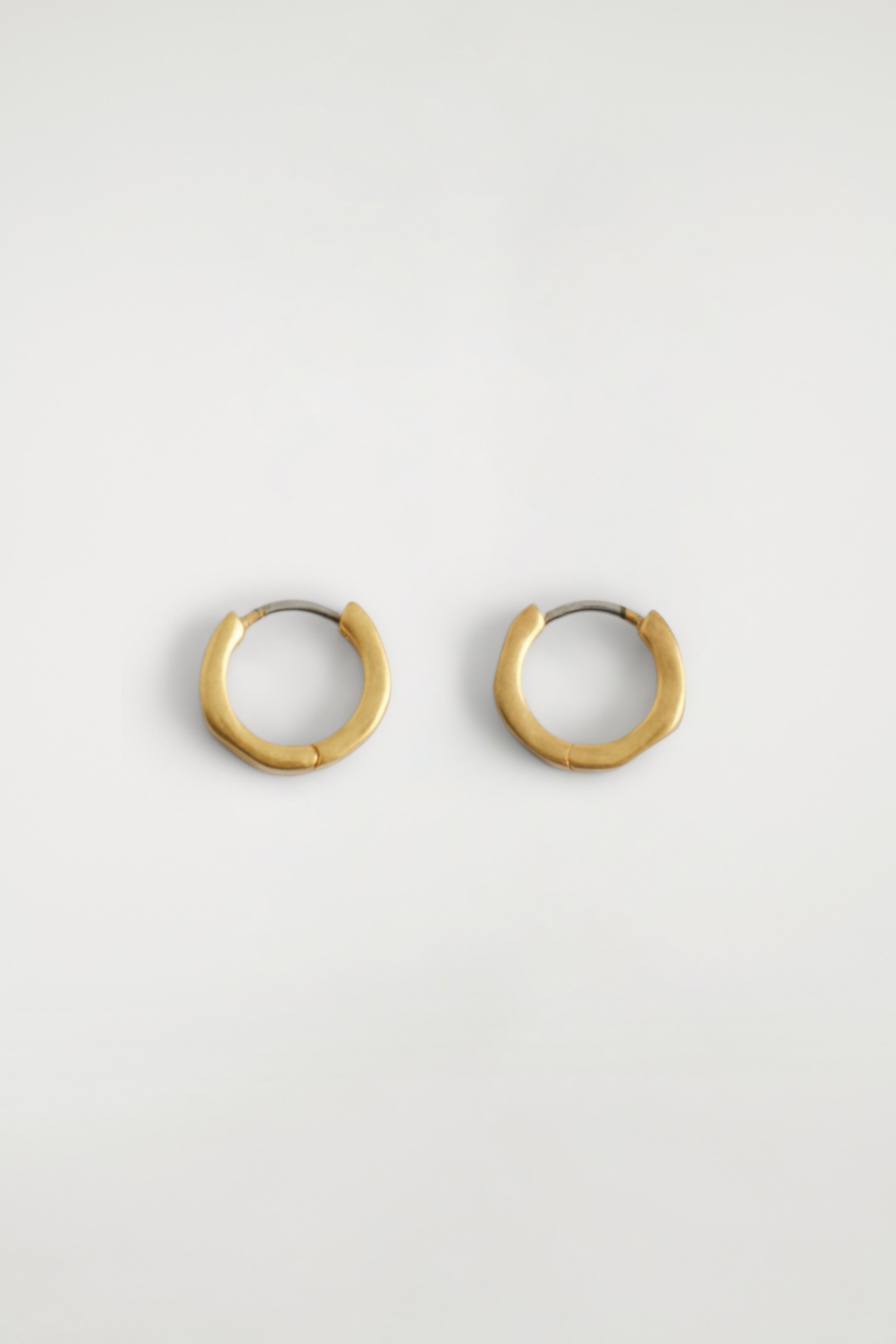 View larger image: Two gold-toned, hammered mini hoop earrings are being presented side-by-side. Each earring features a textured surface with subtle indentations, and a darker metal hinge post is visible at its top.