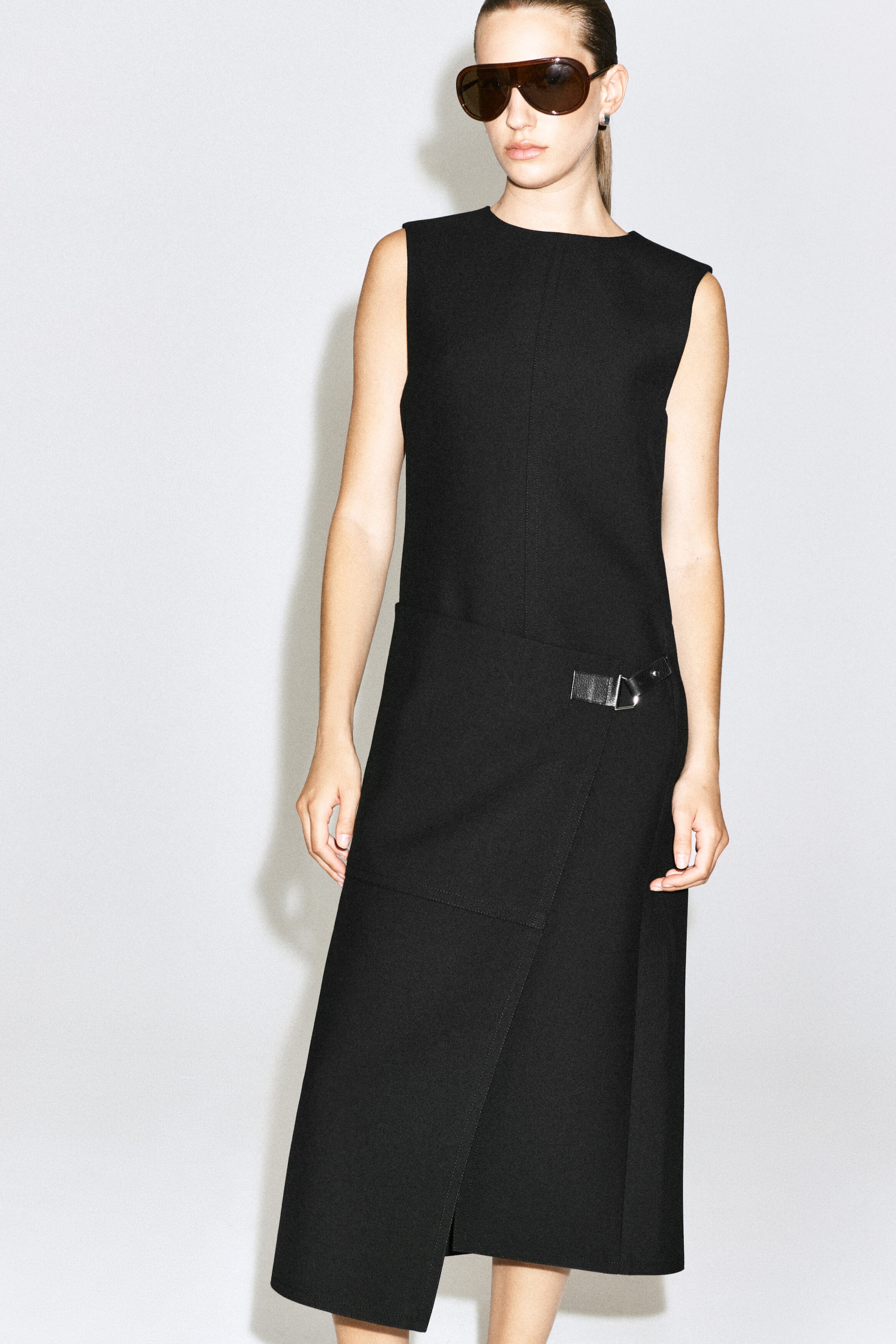 The model is wearing the black belt-detail double-faced wool midi dress. This sleeveless dress features a high, round neckline and a structured, asymmetrical wrap-style skirt accented with a small leather strap and buckle at the hip.