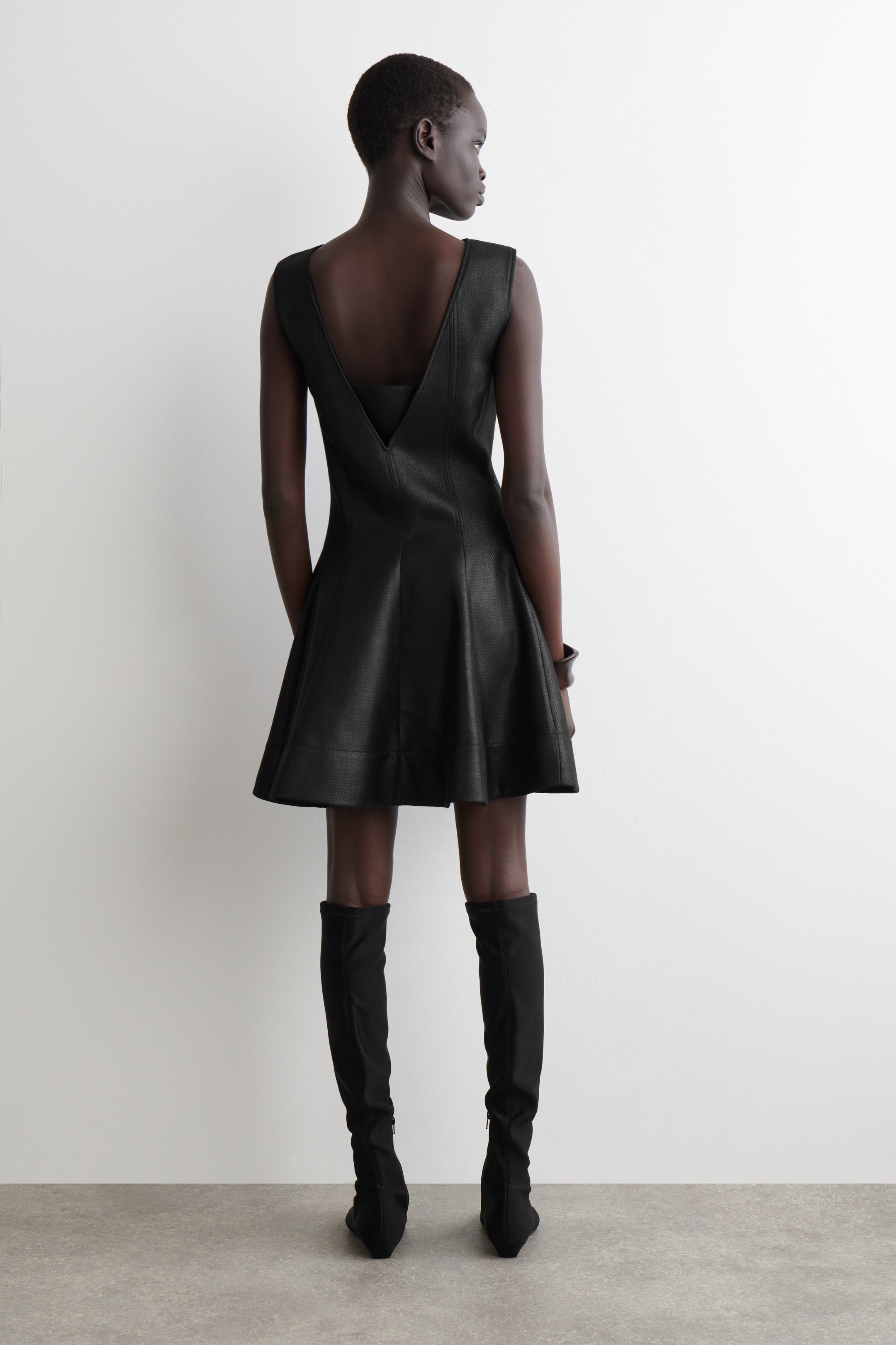 View larger image: A black sleeveless dress is being presented from the back, featuring a deep V-backline with a visible inner band, a fitted bodice, and a flared, above-the-knee skirt made from a textured woven material. Black knee-high boots are being worn with the dress.