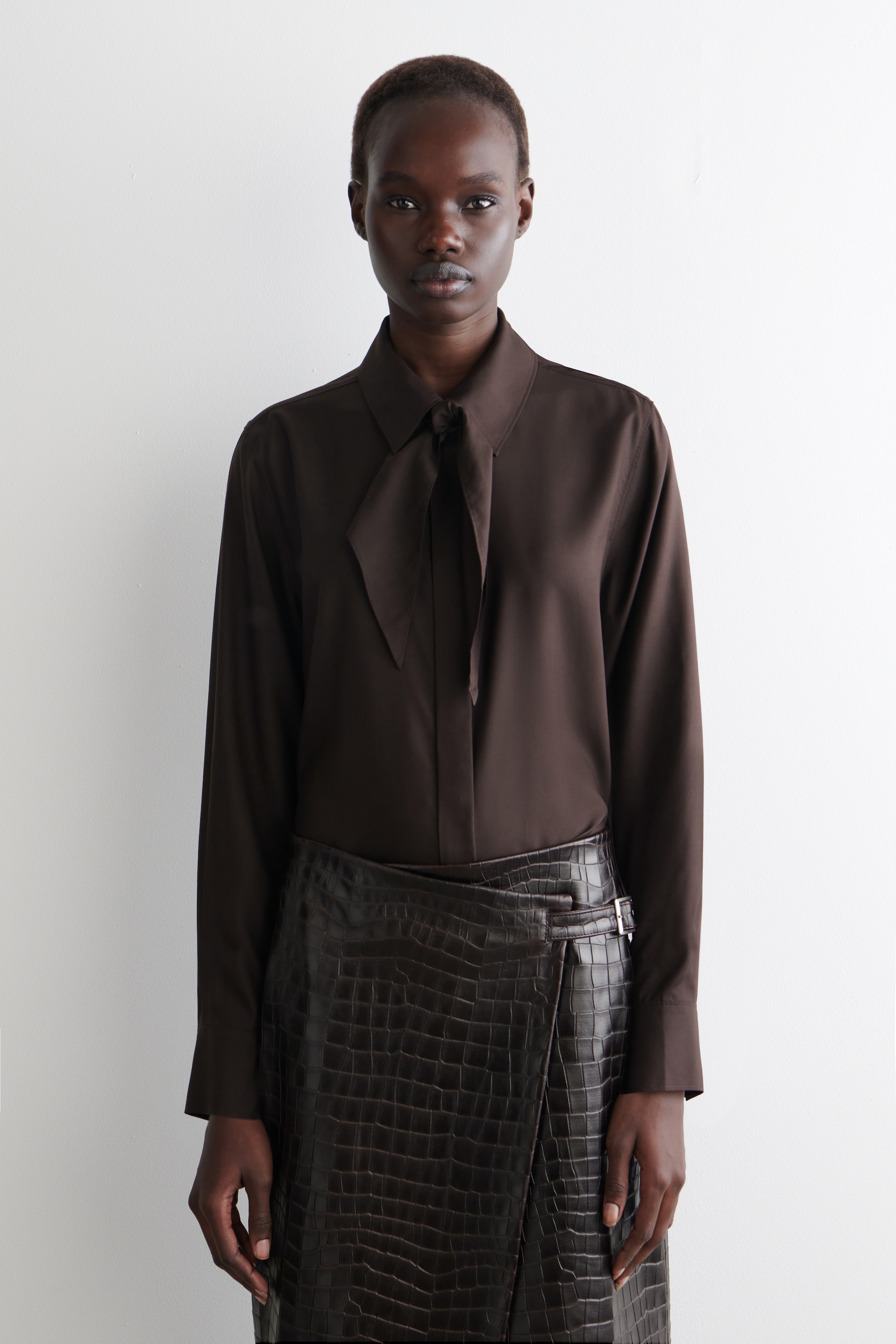 Ver imagen más grande: Una blusa marrón oscuro se presenta de frente, con un cuello en punta y un detalle de lazo en el cuello que está anudado en el centro. La blusa se combina con una falda envolvente marrón oscuro, que muestra una textura de cocodrilo en relieve y una hebilla plateada en la cadera izquierda.