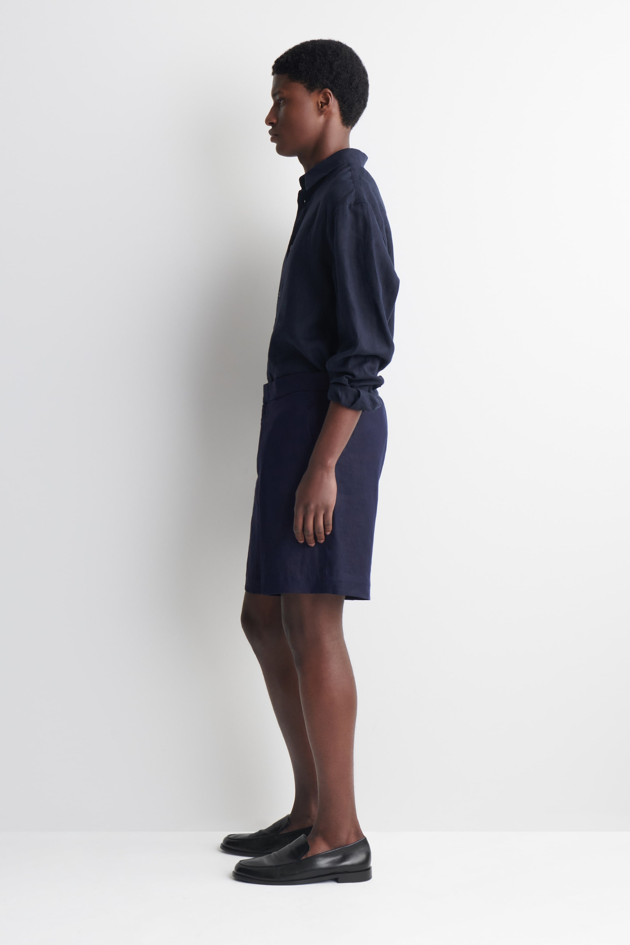 Visualizza immagine più grande: Una camicia di lino blu scuro con bottoni, con le maniche lunghe arrotolate, e pantaloncini coordinati sono presentati in un profilo laterale. Mocassini neri completano l'outfit.