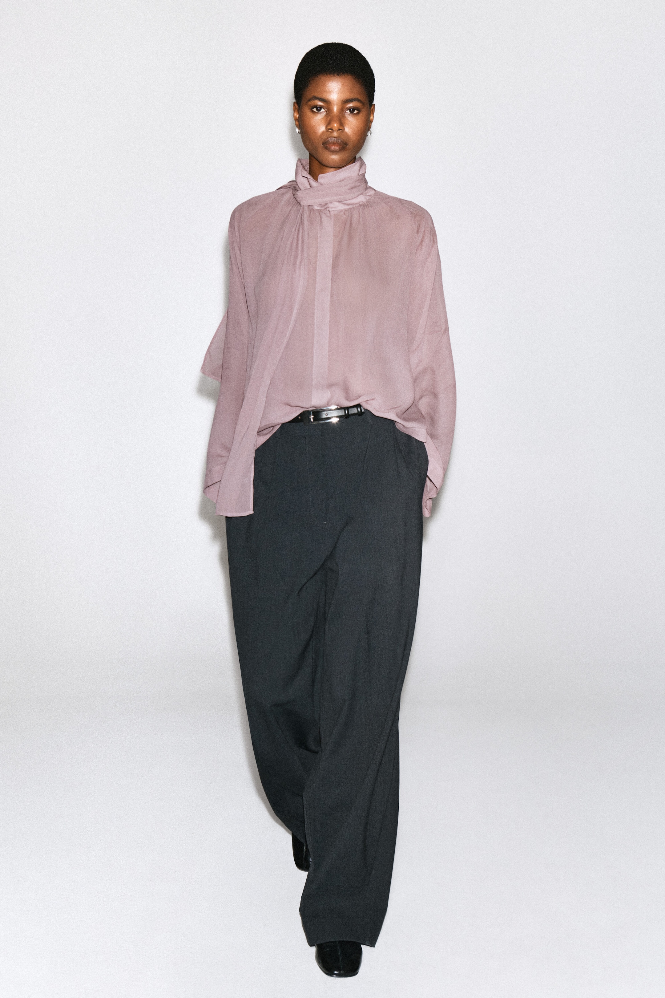 Un haut mauve fluide et semi-transparent, doté d’un col montant avec détail écharpe intégré, est porté par le mannequin. La blouse à manches longues est stylisée avec un rentré partiel dans un pantalon foncé à jambes larges.