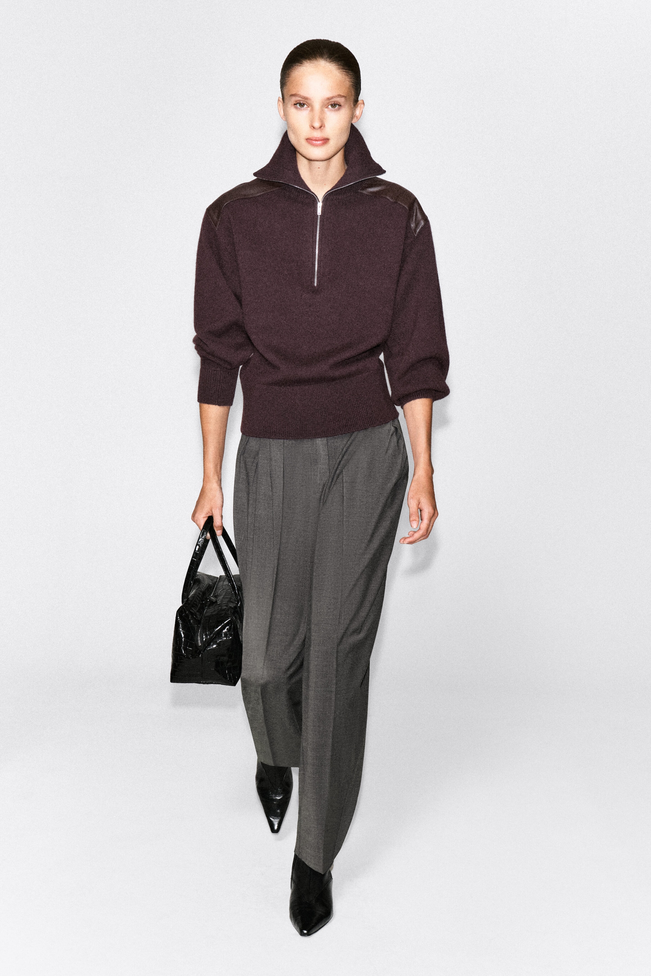 The model is wearing a dark plum leather-patch merino wool zip-up jumper featuring a high collar, a silver half-zip fastening, and leather panels on the shoulders. The jumper is styled with gray wide-leg trousers, black pointed-toe boots, and a black leather handbag. The model is wearing a dark plum leather-patch merino wool zip-up jumper featuring a high collar, a silver half-zip fastening, and leather panels on the shoulders. The jumper is styled with gray wide-leg trousers, black pointed-toe boots, and a black leather handbag.