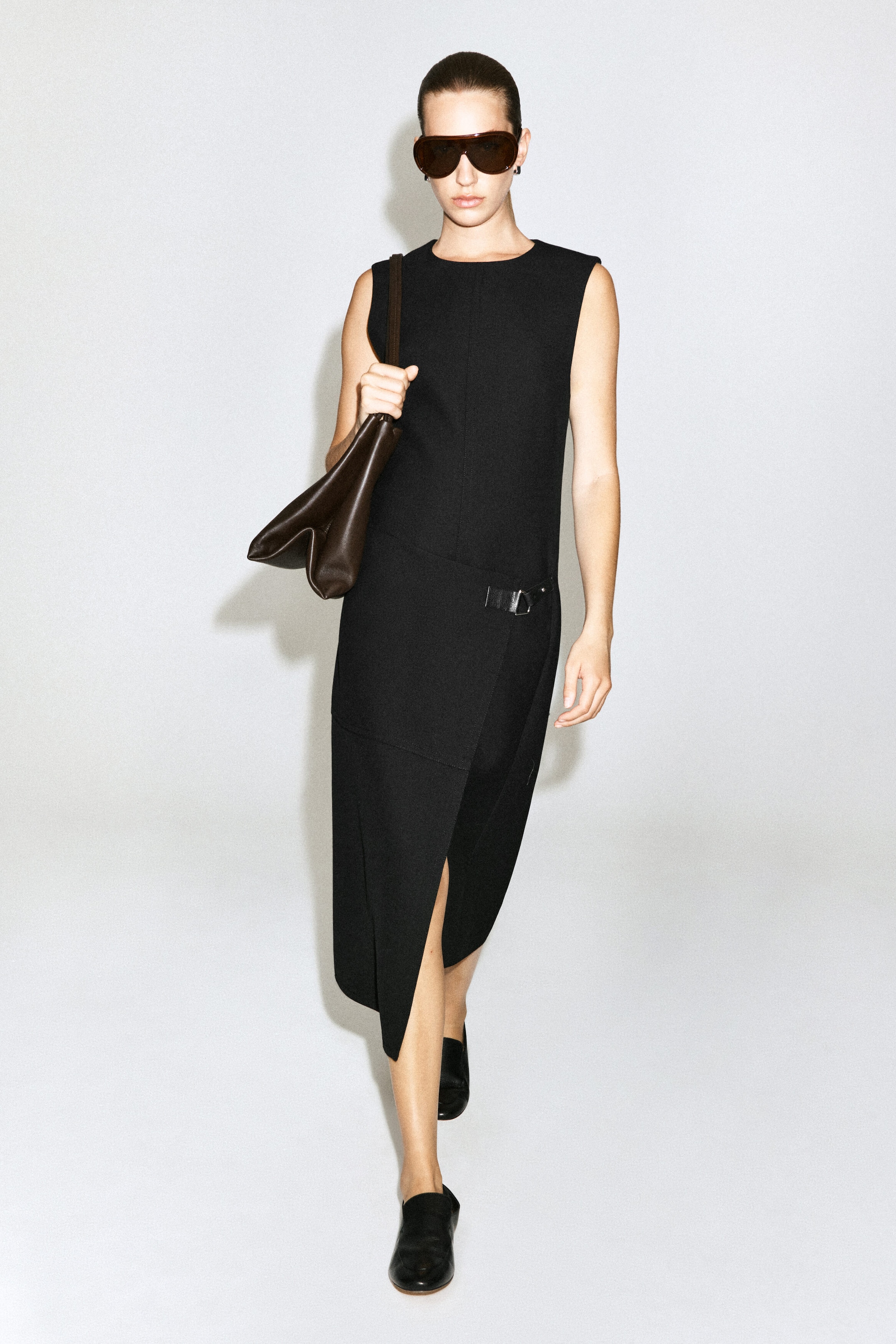 The black belt-detail double-faced wool midi dress is worn by the model and features a sleeveless design, a round neckline, and a wrap-style skirt with a front slit and a small buckle detail at the hip. The dress is styled with a dark brown leather shoulder bag, black loafers, and oversized sunglasses.