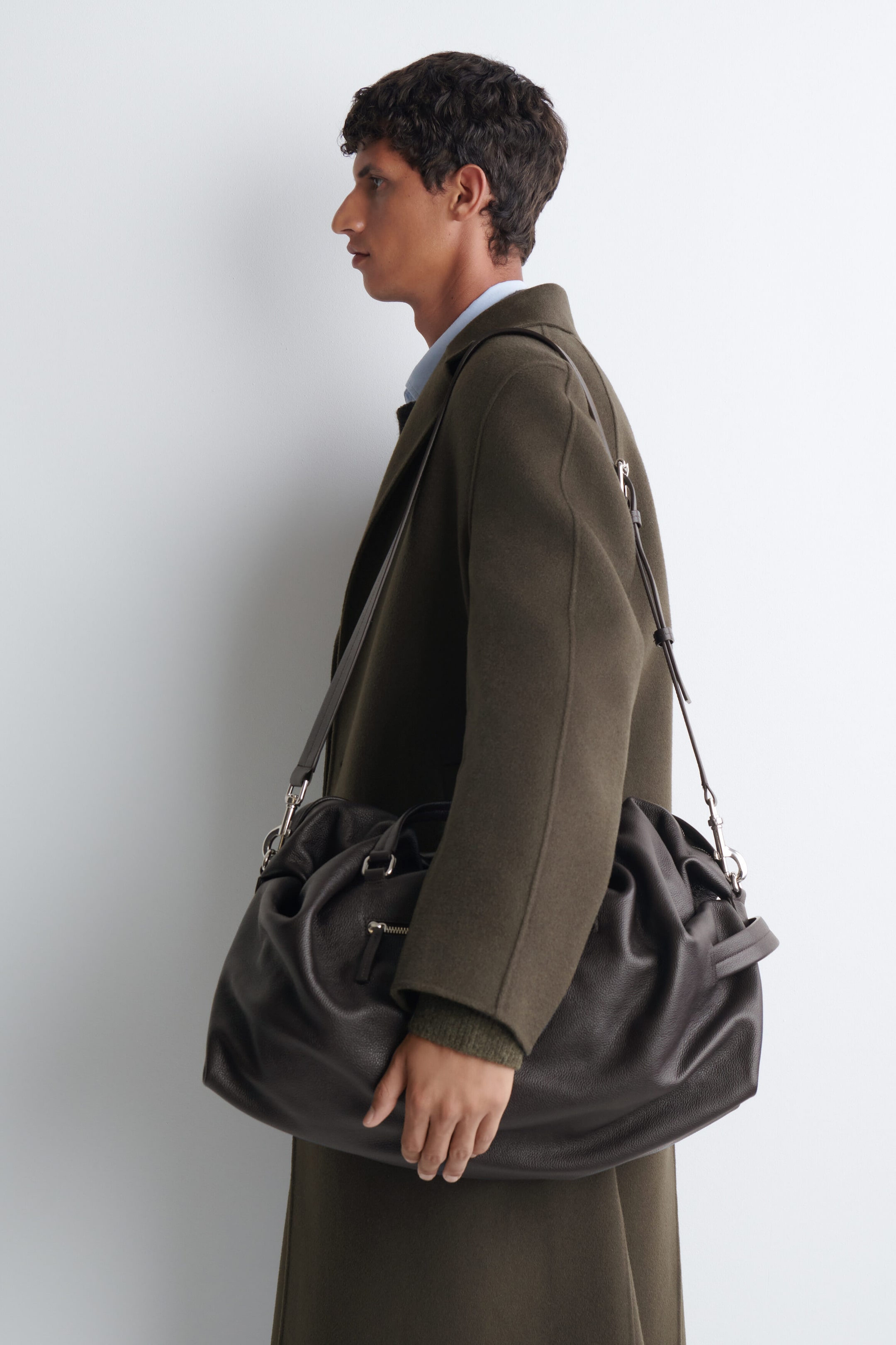 A dark brown pebbled leather duffle bag is being carried over the shoulder of the model, who is wearing an olive green coat. The slouchy bag features dual top handles, a zipped exterior pocket, and a long leather strap resting on the model's shoulder.