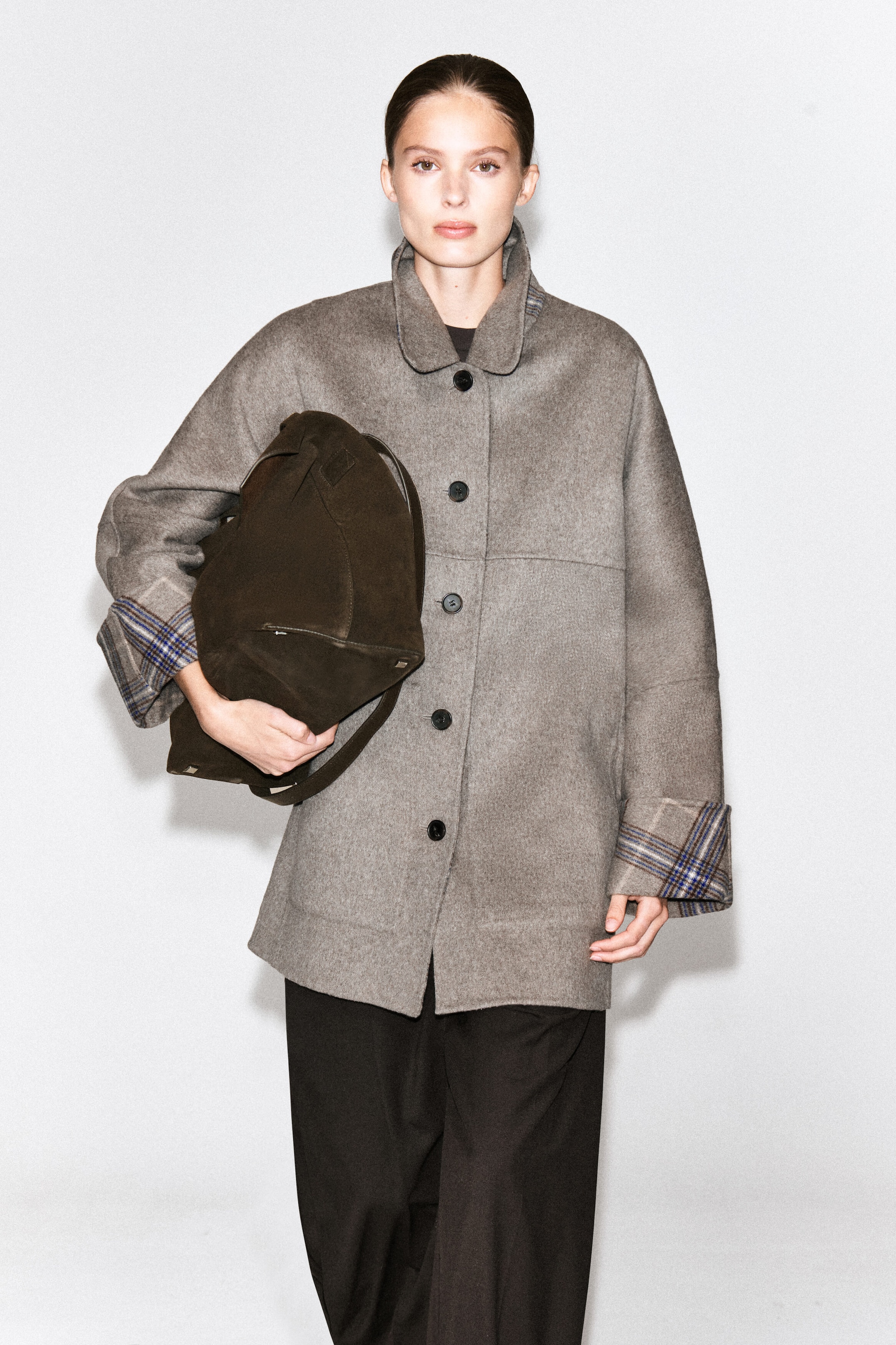 A double-faced wool-blend coat in a muted gray-beige is worn by the model, featuring a button-front closure, a classic collar, and turned-up cuffs that reveal a blue-and-brown check pattern on the reverse. The coat is styled with dark trousers, and the model is carrying a dark brown suede bag under one arm.