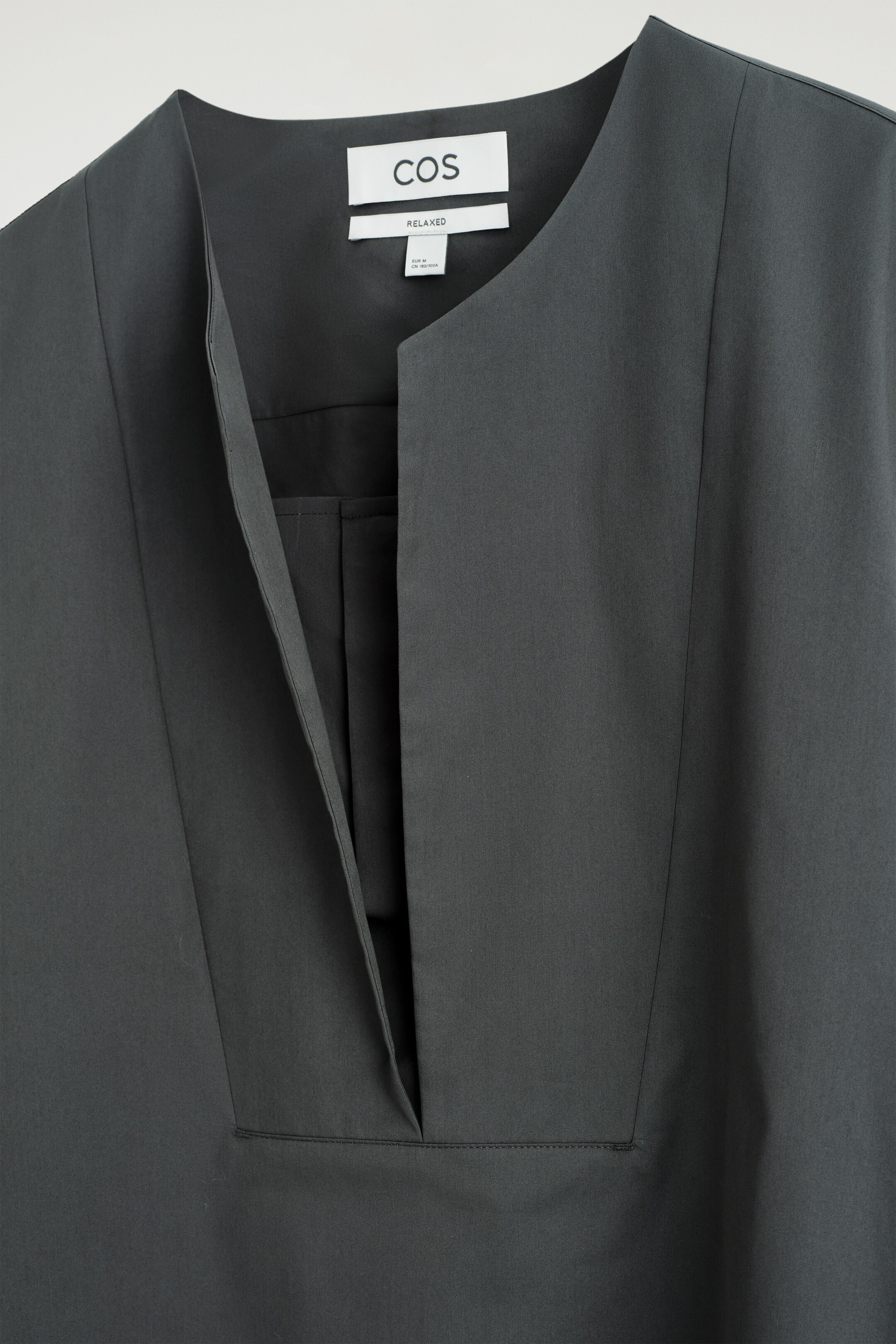 Ver imagen más grande: Se muestra una parte superior de color gris oscuro desde el frente, con un escote en V profundo y angular que forma una abertura estructurada. Una etiqueta blanca de la marca COS, que indica ajuste "RELAXED", es visible en la parte posterior del cuello.