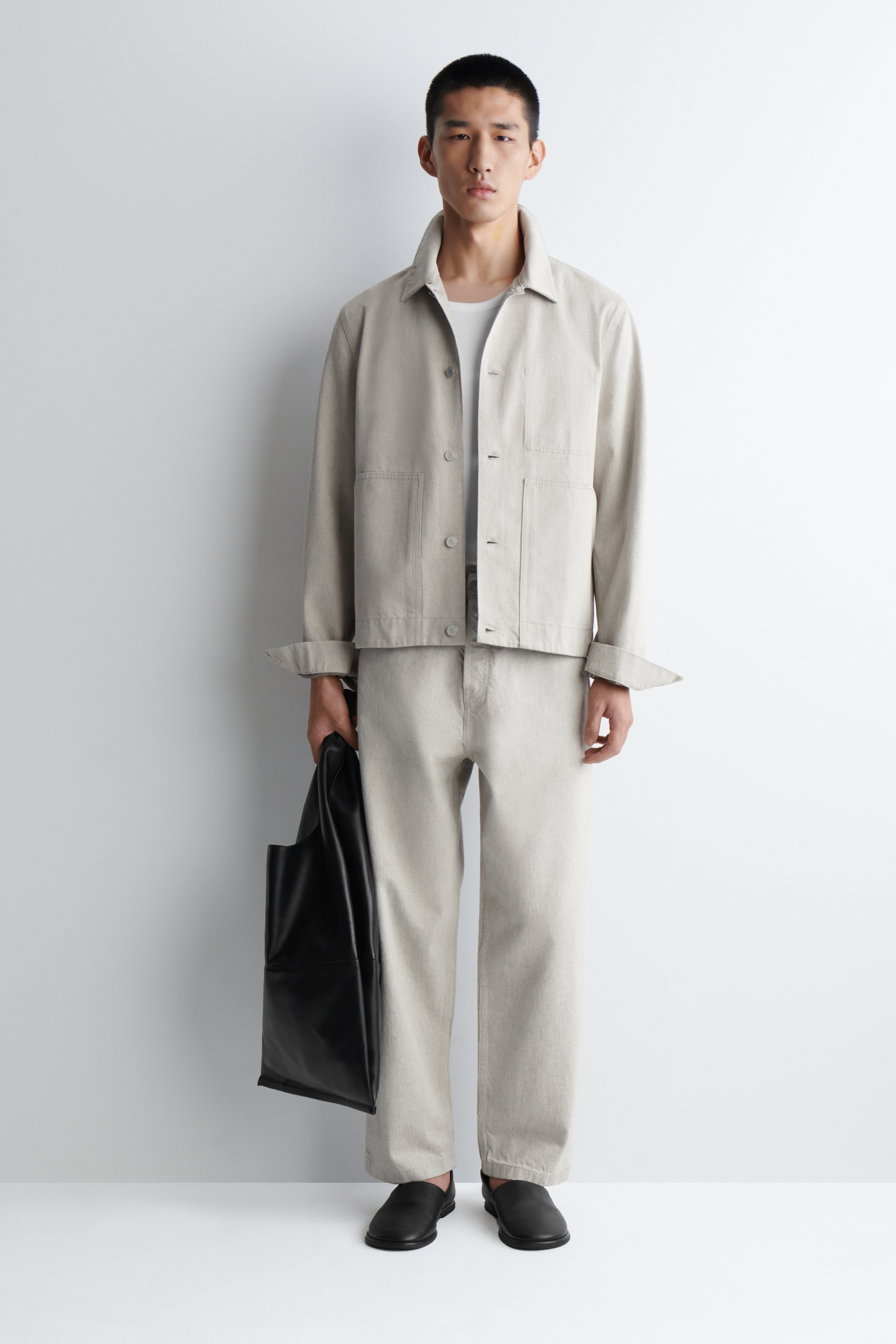 Visualizza immagine più grande: Una giacca da lavoro beige chiaro è indossata sopra una t-shirt bianca con scollo a giro, abbinata a pantaloni a gamba dritta a vestibilità rilassata coordinati. La giacca presenta un colletto a punta, chiusura frontale con bottoni e quattro tasche applicate, con i polsini arrotolati, mentre scarpe nere slip-on e una borsa tote nera completano l'ensemble.