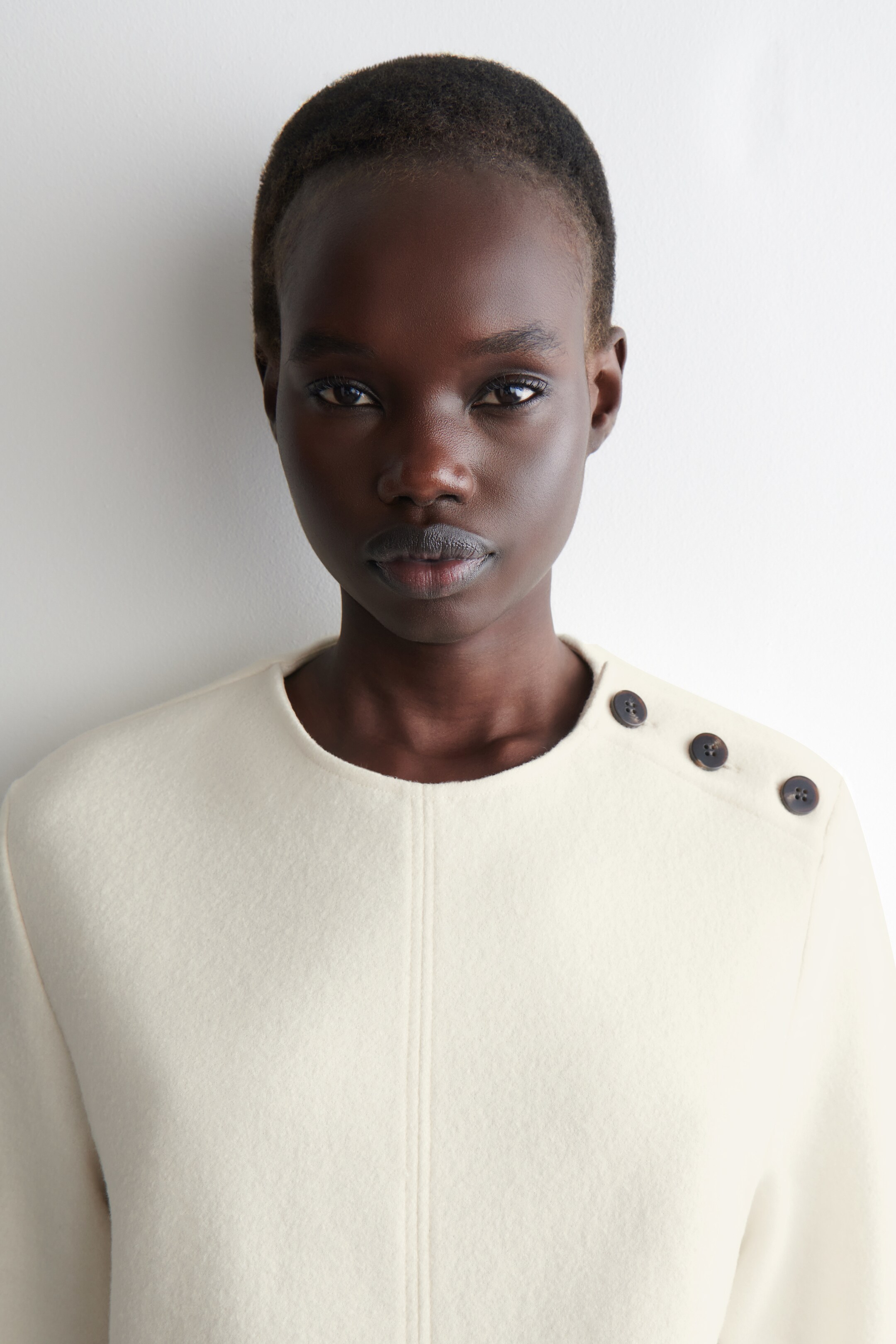 Agrandir l'image: Un haut ou une veste blanc cassé est présenté de face, avec un col rond et une couture verticale centrale. Trois boutons ronds et sombres ornent l'épaule droite, apparaissant comme un détail décoratif.