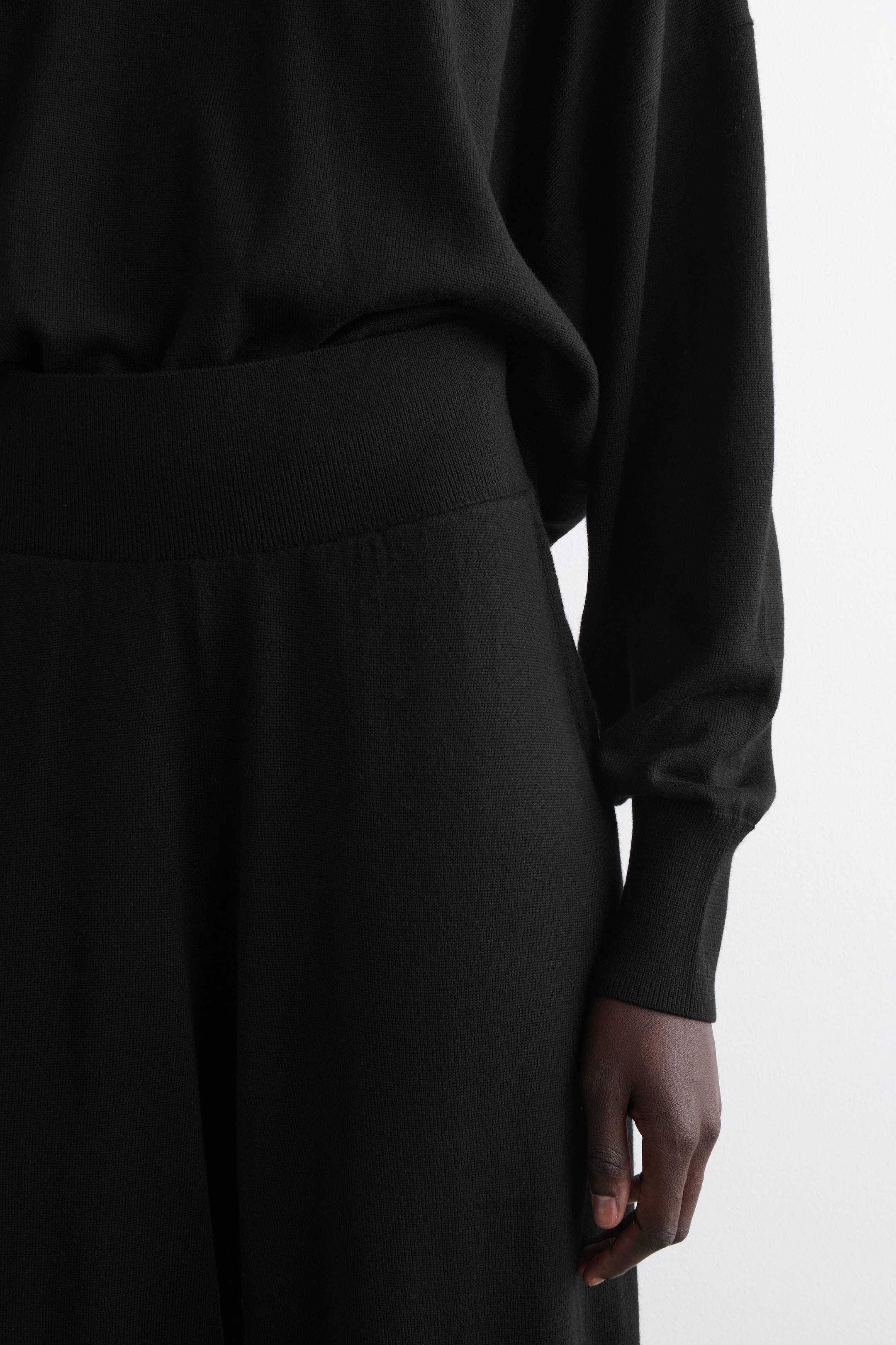Visualizza immagine più grande: Un completo nero in maglia a due pezzi viene presentato da un'angolazione leggermente laterale. La parte superiore presenta una vestibilità ampia con maniche lunghe e polsini a coste, e il suo orlo a coste si appoggia sopra l'ampia fascia in vita a coste del fondo coordinato. Il fondo è realizzato in un tessuto a maglia liscia che cade verso il basso.