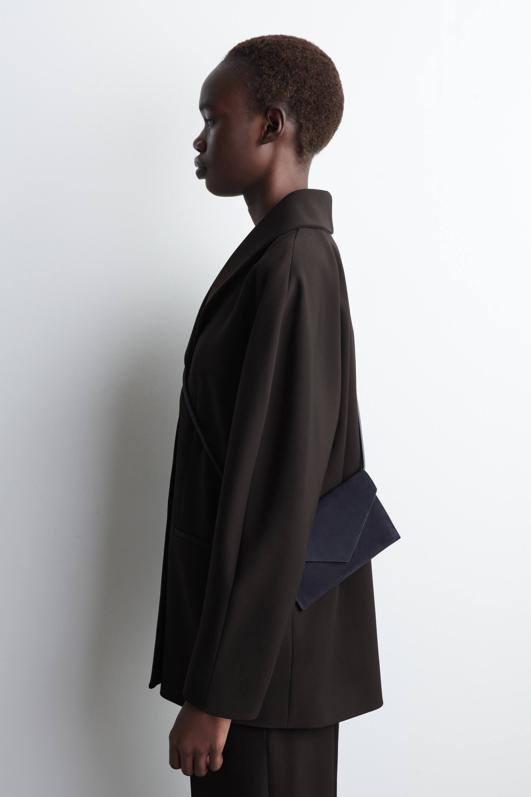 Grösseres Bild ansehen: Ein dunkelbrauner Blazer mit entspannter Passform wird getragen, der über ein breites Revers und lange Ärmel verfügt und aus einer Seitenansicht zu sehen ist. Eine dunkelblaue Umhängetasche im Kuvert-Stil mit einem dünnen Riemen ruht auf der Hüfte des Trägers und wird über dem Blazer getragen.