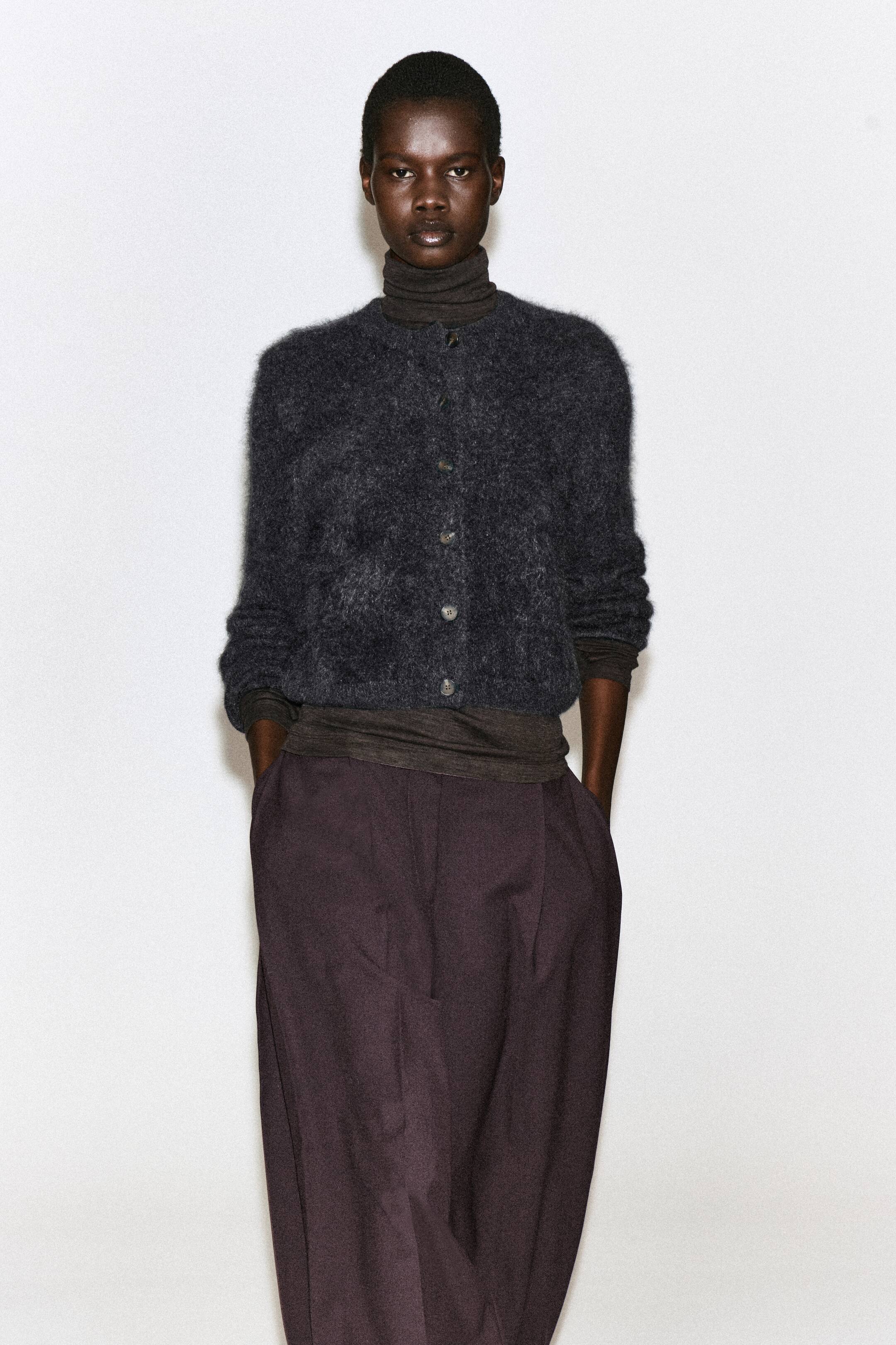 Visualizza l'immagine ingrandita: Un cardigan girocollo in puro cashmere spazzolato, in grigio scuro, indossato abbottonato dalla modella. Il cardigan dalla texture morbida e soffice è sovrapposto a un top dolcevita marrone scuro ed è abbinato a pantaloni scuri a gamba larga.