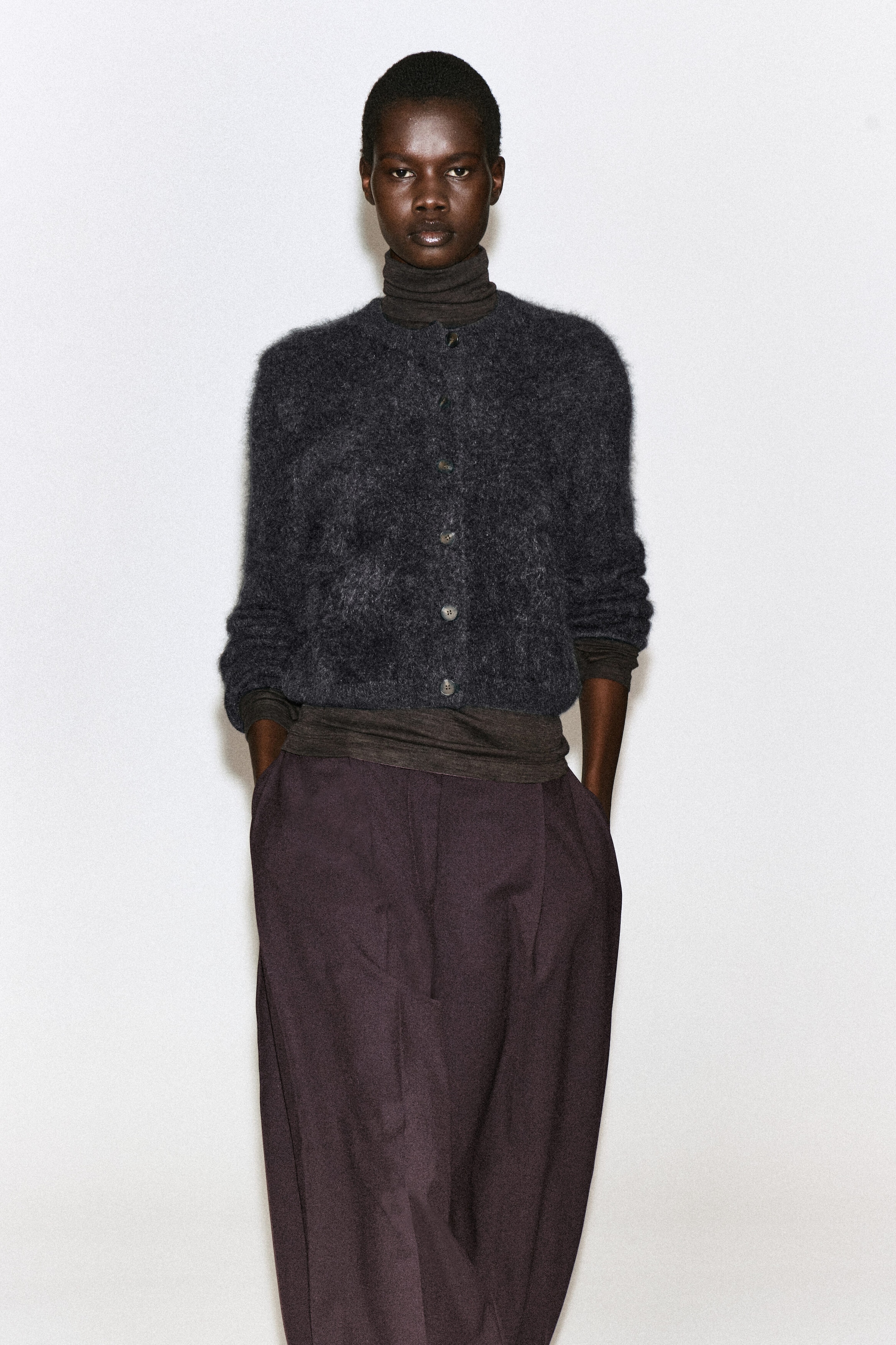 A brushed pure cashmere crew-neck cardigan in dark grey is being worn buttoned up by the model. The fuzzy-textured cardigan is layered over a dark brown turtleneck top and styled with dark, wide-leg trousers. A brushed pure cashmere crew-neck cardigan in dark grey is being worn buttoned up by the model. The fuzzy-textured cardigan is layered over a dark brown turtleneck top and styled with dark, wide-leg trousers.