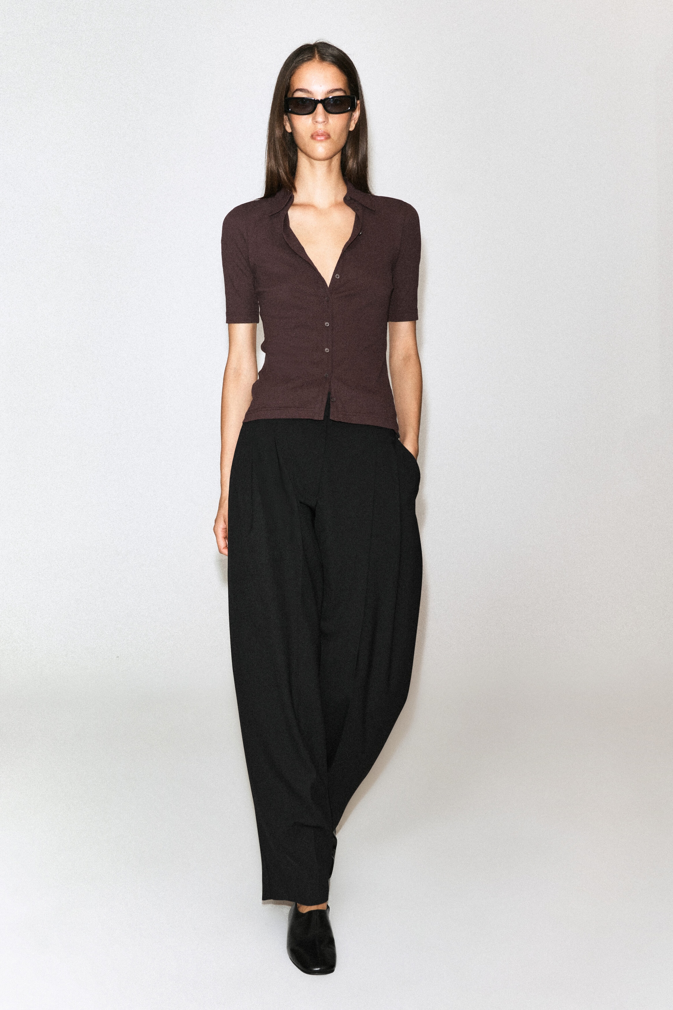 A dark brown, short-sleeved sculpted cotton jersey shirt featuring a classic collar, button-front closure, and fitted silhouette is worn by the model. The shirt is styled untucked over wide-leg black trousers. A dark brown, short-sleeved sculpted cotton jersey shirt featuring a classic collar, button-front closure, and fitted silhouette is worn by the model. The shirt is styled untucked over wide-leg black trousers.