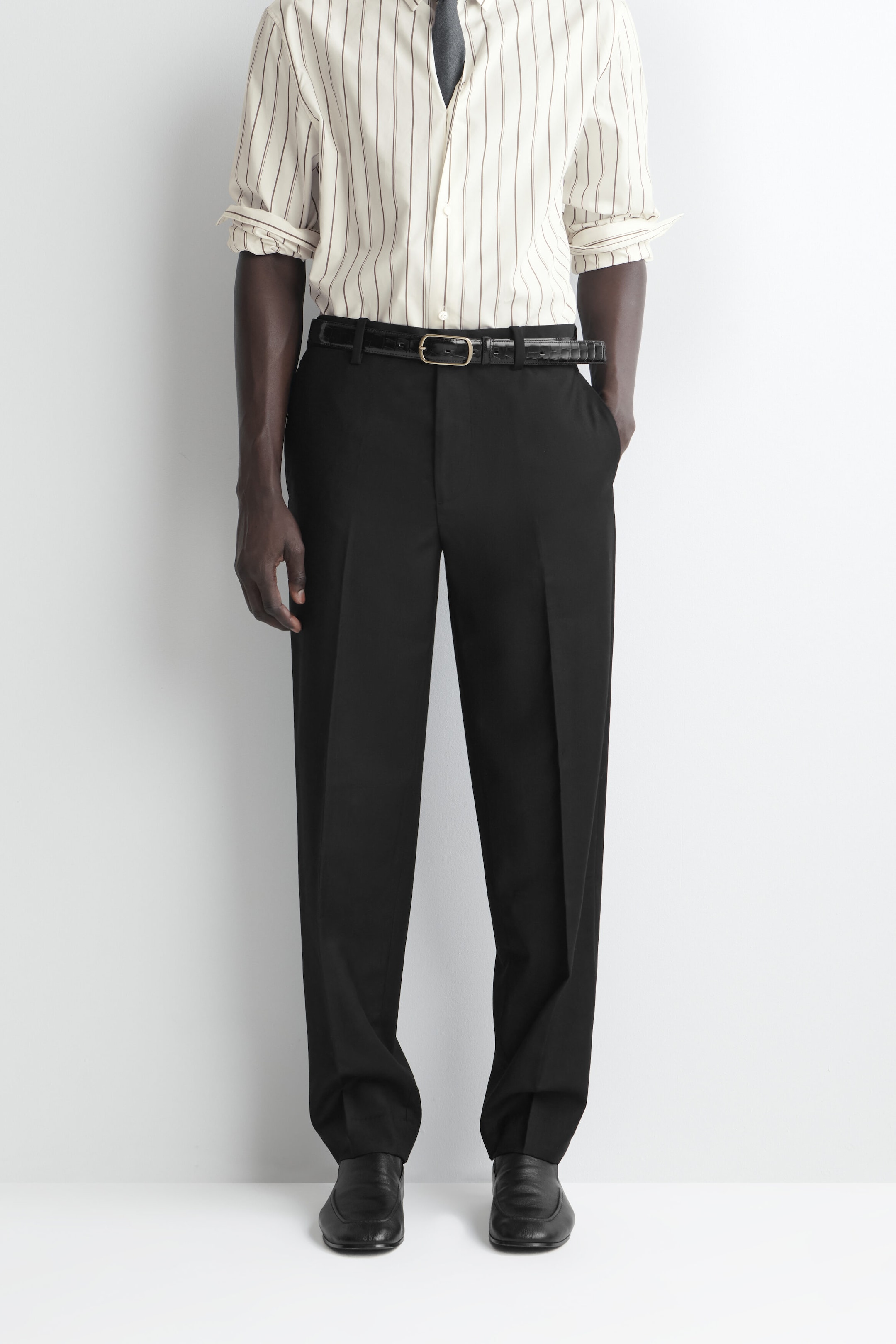 These black straight-leg trousers are shown from the waist down, featuring a regular fit with sharp pressed creases running down the centre of each leg. They are styled with a black textured belt and a tucked-in striped shirt, draping straight down to pool slightly over black leather shoes. These black straight-leg trousers are shown from the waist down, featuring a regular fit with sharp pressed creases running down the centre of each leg. They are styled with a black textured belt and a tucked-in striped shirt, draping straight down to pool slightly over black leather shoes.
