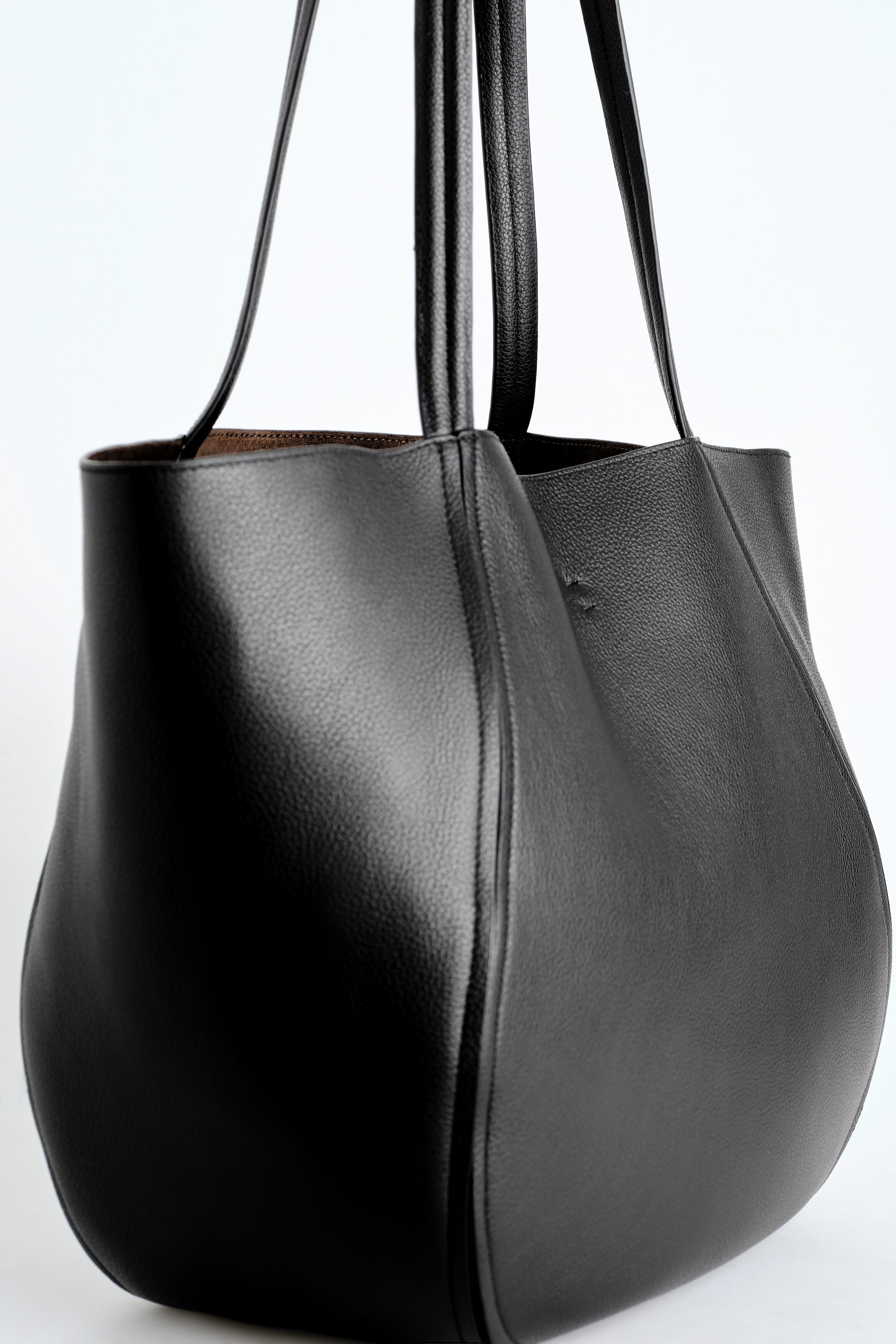A black grained leather shopper bag is facing left, showcasing its softly curved silhouette, vertical pleated seams, and textured surface. Two long, slim shoulder straps extend upward, while the open top reveals a glimpse of the contrasting brown interior lining. A black grained leather shopper bag is facing left, showcasing its softly curved silhouette, vertical pleated seams, and textured surface. Two long, slim shoulder straps extend upward, while the open top reveals a glimpse of the contrasting brown interior lining.