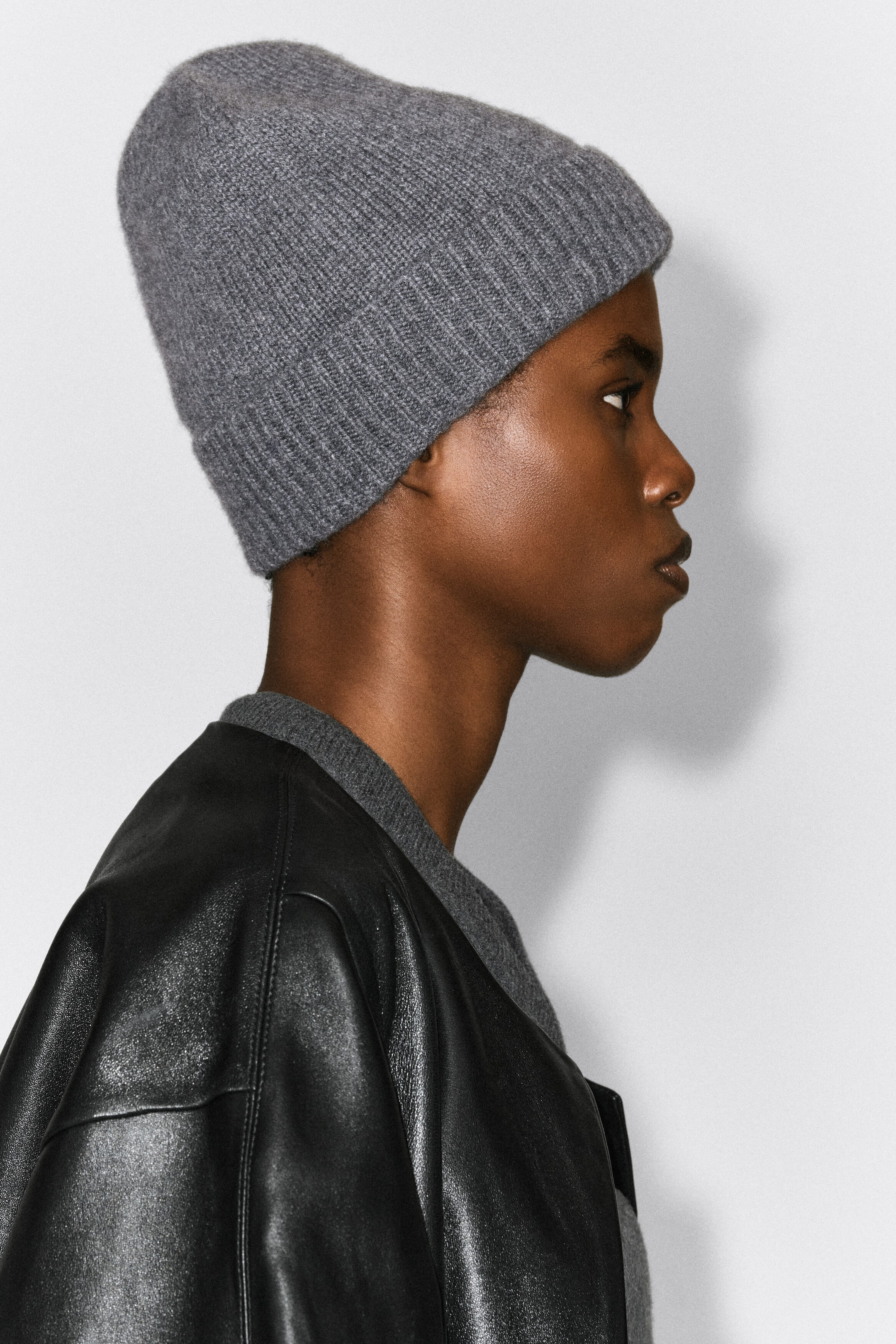 A grey cashmere beanie featuring a ribbed cuff is being worn by the model, who is shown in profile. The beanie is styled alongside a coordinating grey knit top and a black leather jacket. A grey cashmere beanie featuring a ribbed cuff is being worn by the model, who is shown in profile. The beanie is styled alongside a coordinating grey knit top and a black leather jacket.