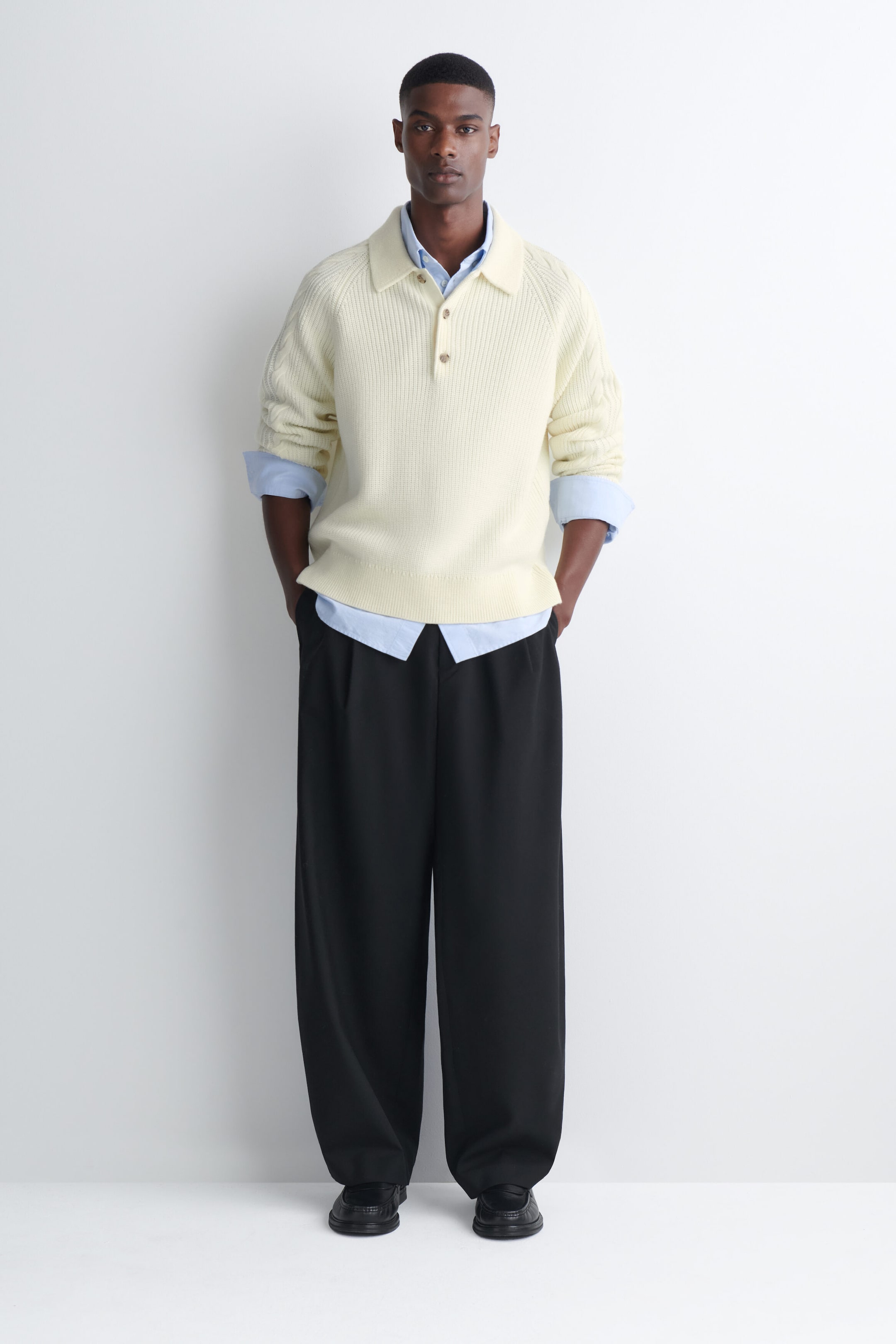 A cream-colored chunky knit polo sweater featuring a collar, button placket, and cable-knit sleeve details is being worn by the model. The sweater is layered over a light blue button-down shirt and styled with wide-leg black trousers. A cream-colored chunky knit polo sweater featuring a collar, button placket, and cable-knit sleeve details is being worn by the model. The sweater is layered over a light blue button-down shirt and styled with wide-leg black trousers.