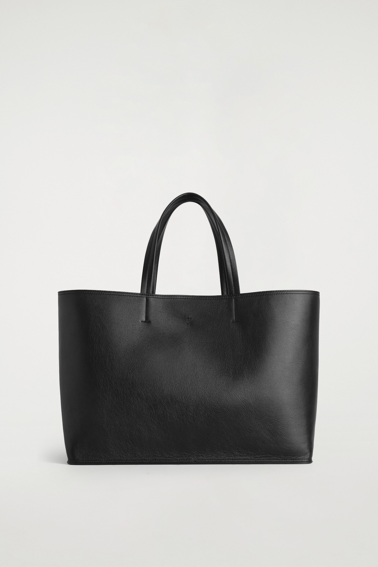 A black pebbled leather tote bag stands upright facing forward, featuring a structured rectangular silhouette and two rounded top handles attached with tonal stitching.