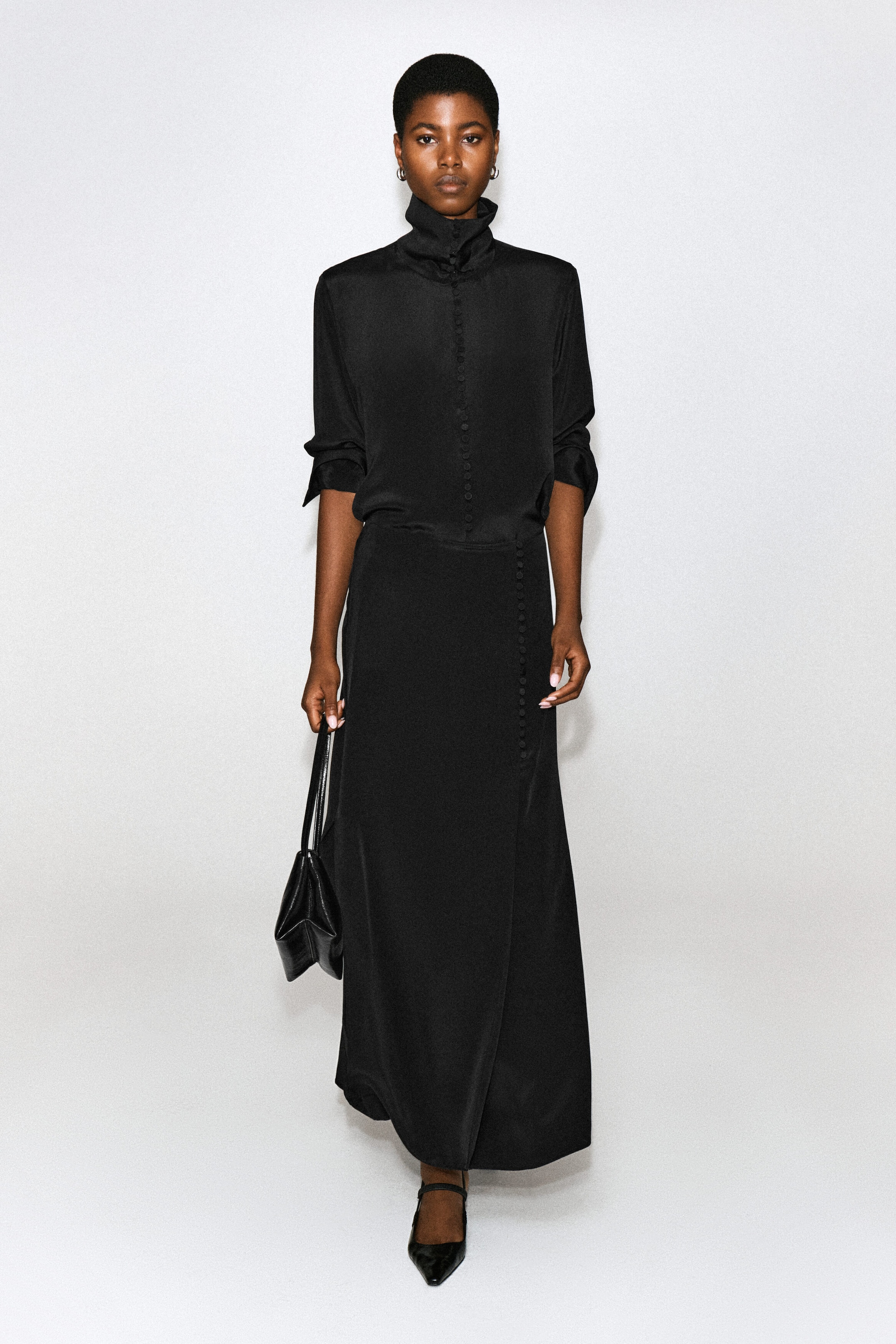 The model is wearing a black high-neck long-sleeve blouse with front button detailing and a matching maxi skirt. She is carrying a small black leather handbag in one hand and wearing black pointed-toe shoes.