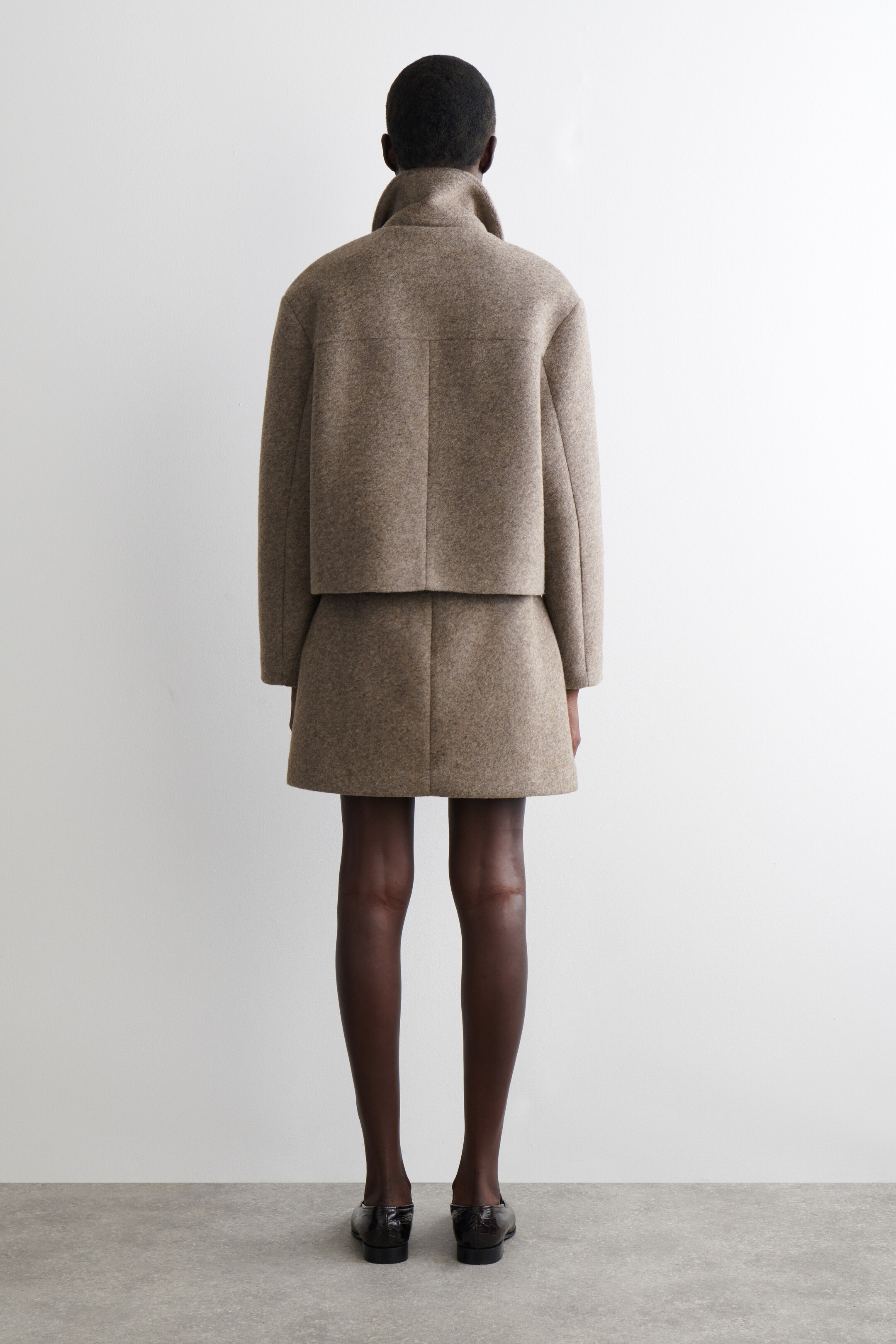 Visualizza immagine più grande: Un completo marrone mélange, composto da una giacca corta e una minigonna a trapezio, viene mostrato da dietro. La giacca presenta un colletto alto, un giogo orizzontale sul retro e una cucitura centrale verticale sul retro, mentre la gonna presenta anch'essa una cucitura centrale verticale sul retro.