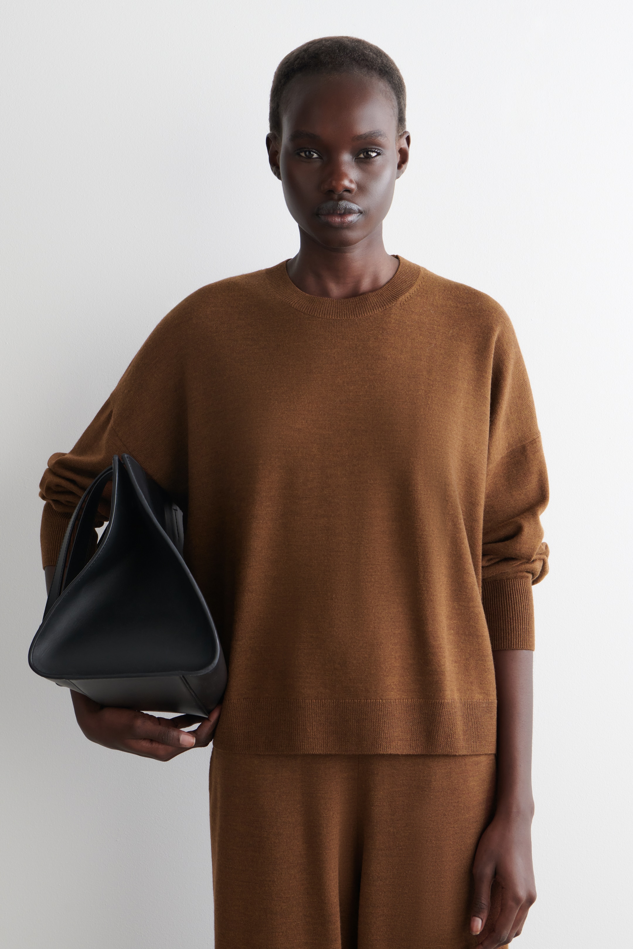 Agrandir l'image: Un ensemble en tricot marron, comprenant un haut à col rond et coupe décontractée avec des poignets et un ourlet côtelés, est présenté. Complétant l'ensemble, un sac structuré noir avec une base triangulaire est tenu sur le côté.