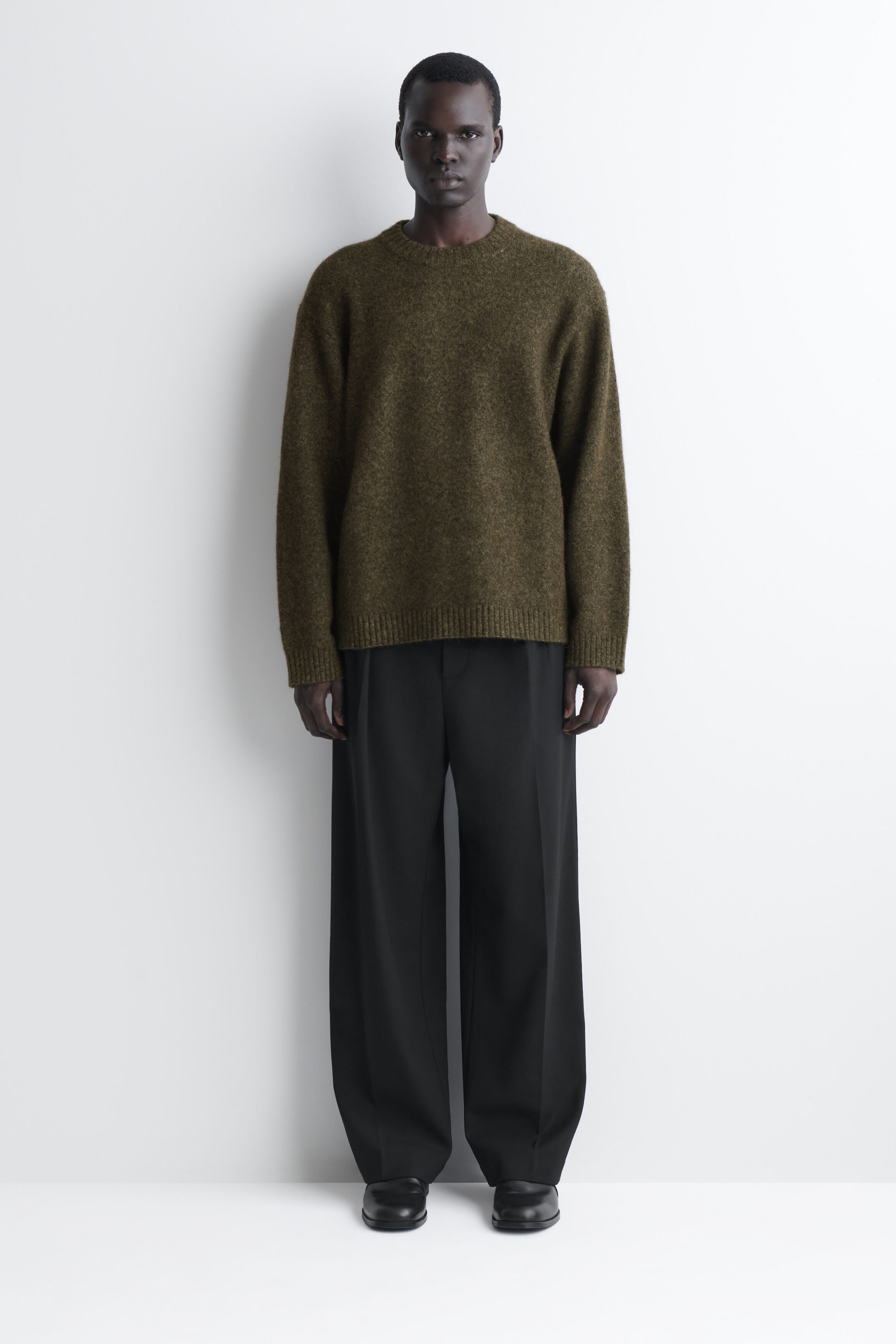 An oversized alpaca-blend crew-neck jumper in a textured dark green knit is shown from the front, highlighting a relaxed fit with dropped shoulders and ribbed detailing at the collar, cuffs, and hem. The jumper is layered over black wide-leg trousers, illustrating its loose silhouette and hip-length cut. An oversized alpaca-blend crew-neck jumper in a textured dark green knit is shown from the front, highlighting a relaxed fit with dropped shoulders and ribbed detailing at the collar, cuffs, and hem. The jumper is layered over black wide-leg trousers, illustrating its loose silhouette and hip-length cut.