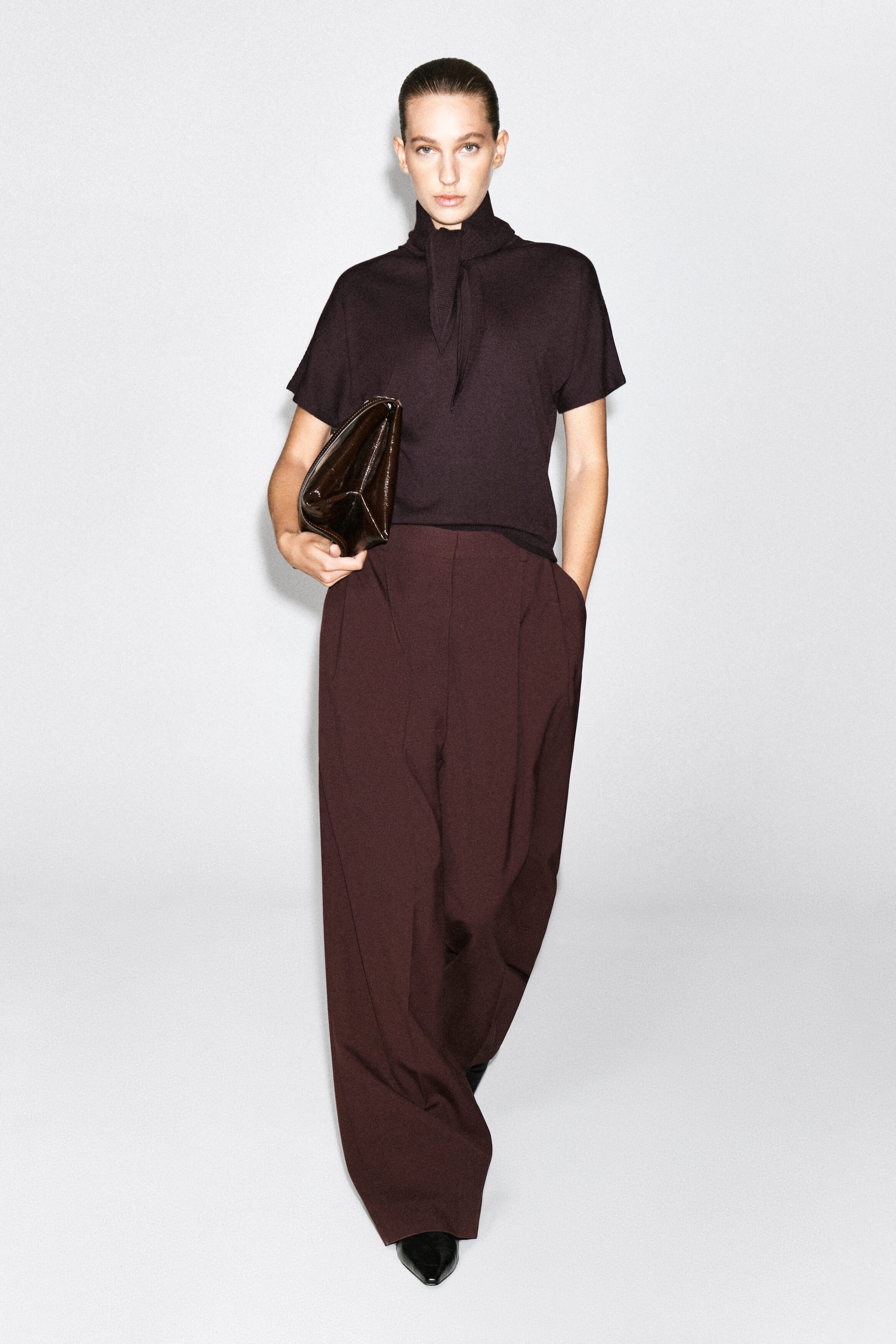 The model is wearing a dark brown merino wool-blend T-shirt with a scarf detail, featuring a high neckline with an integrated knotted scarf detail and short sleeves. The T-shirt is tucked into matching wide-leg trousers, and a glossy dark brown clutch is held under one arm.