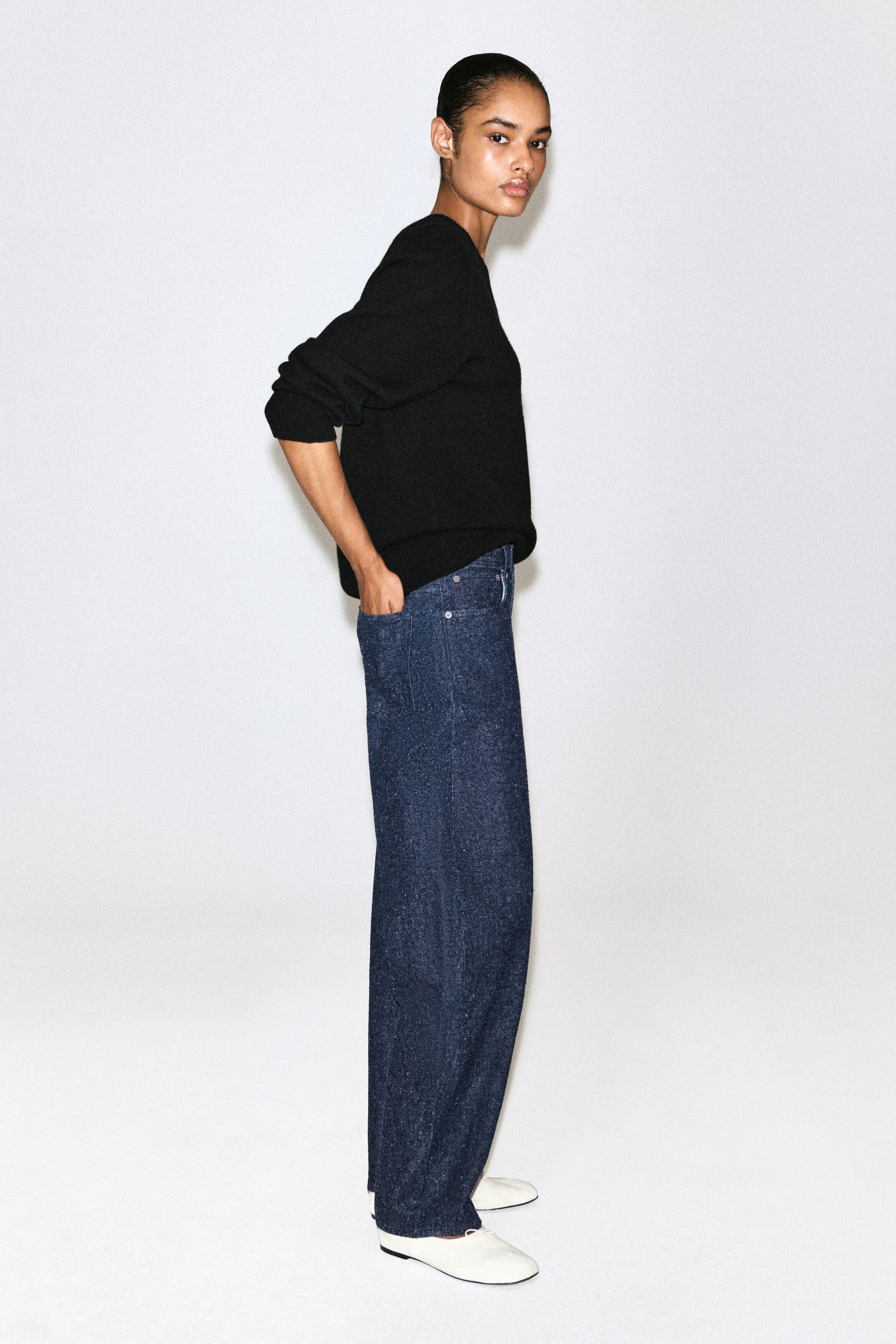 These wide-leg, textured dark blue denim jeans are shown in right-facing profile, highlighting a relaxed straight-leg fit and classic five-pocket styling. The jeans are paired with a black crewneck sweater and white flat slip-on shoes. These wide-leg, textured dark blue denim jeans are shown in right-facing profile, highlighting a relaxed straight-leg fit and classic five-pocket styling. The jeans are paired with a black crewneck sweater and white flat slip-on shoes.