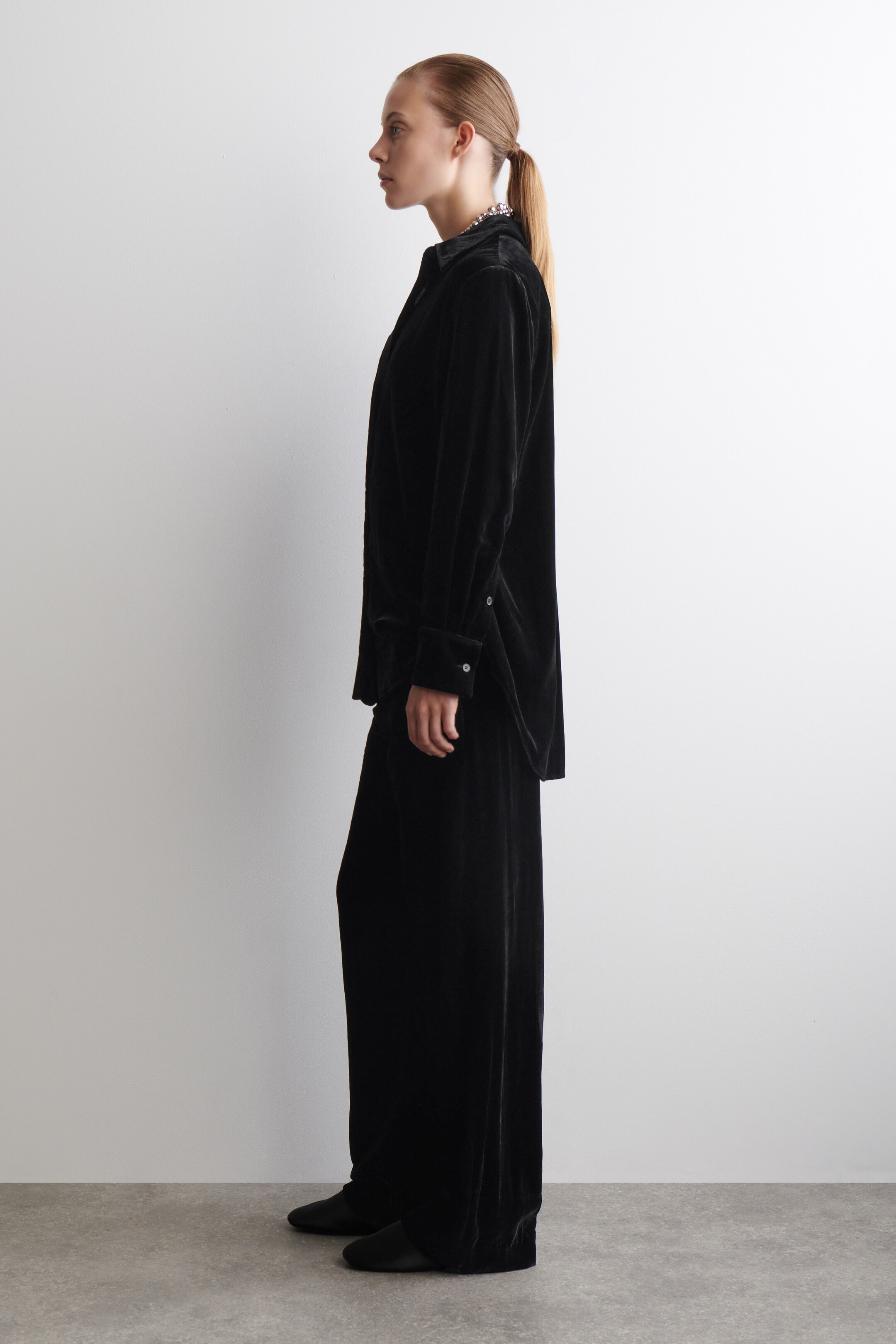 Visualizza immagine più grande: Una camicia nera in velluto con colletto e pantaloni coordinati a gamba larga sono presentati in vista laterale di profilo. La camicia presenta maniche lunghe con polsini con bottoni e una collana con perline è visibile sotto il colletto. I pantaloni sono lunghi fino al pavimento e hanno una vestibilità rilassata.