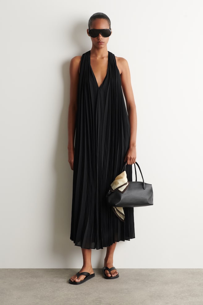 Das schwarze plissierte Kleid ist nach vorne gerichtet und zeigt einen Neckholder-Ausschnitt sowie einen tiefen V-Ausschnitt, mit durchgehenden vertikalen Falten, die in eine Midi-Länge fließen. - 1