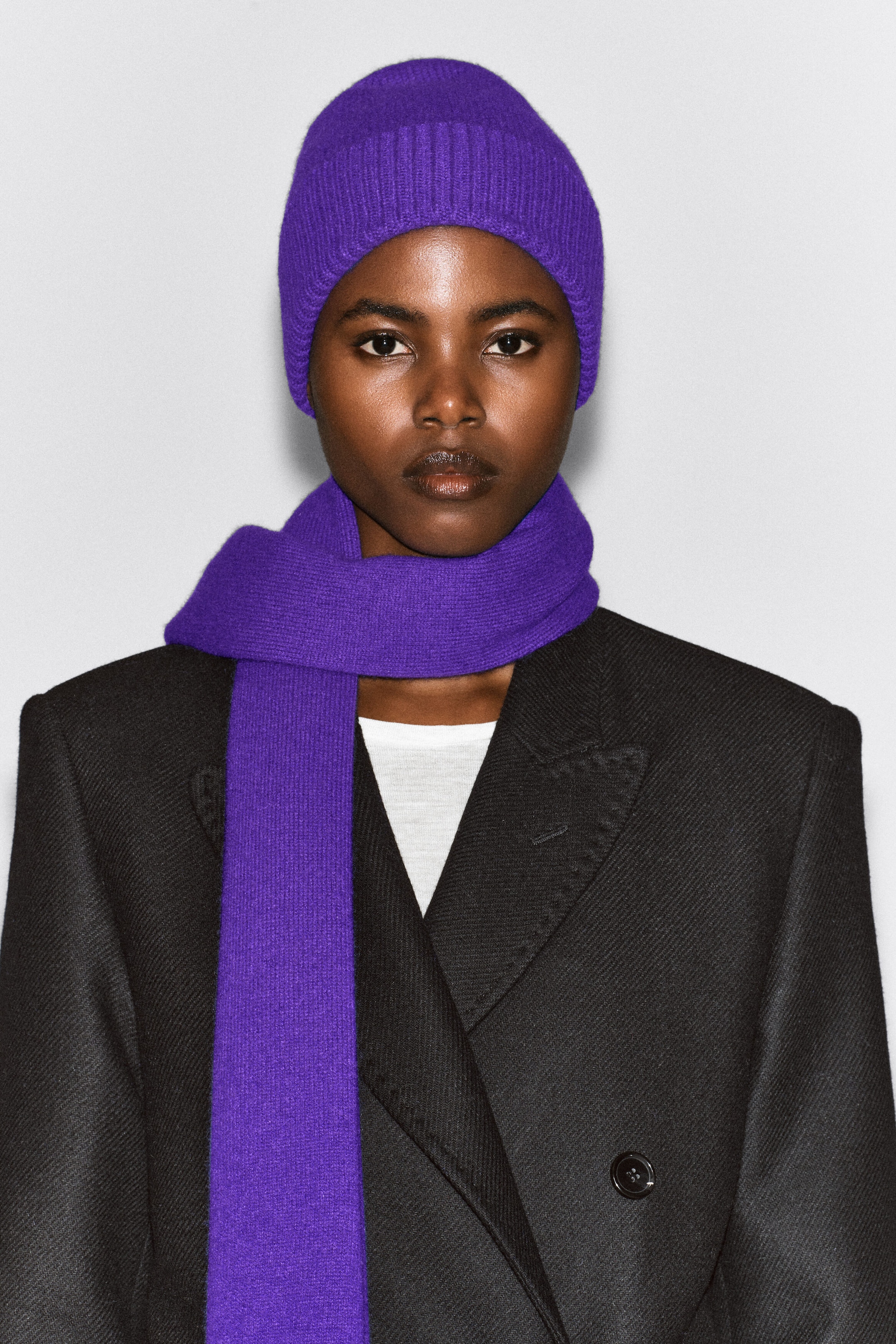 A purple knit cashmere scarf is worn wrapped once around the model’s neck, with one long end hanging down the front over a black double-breasted coat. The scarf is paired with a matching purple knit beanie. A purple knit cashmere scarf is worn wrapped once around the model’s neck, with one long end hanging down the front over a black double-breasted coat. The scarf is paired with a matching purple knit beanie.