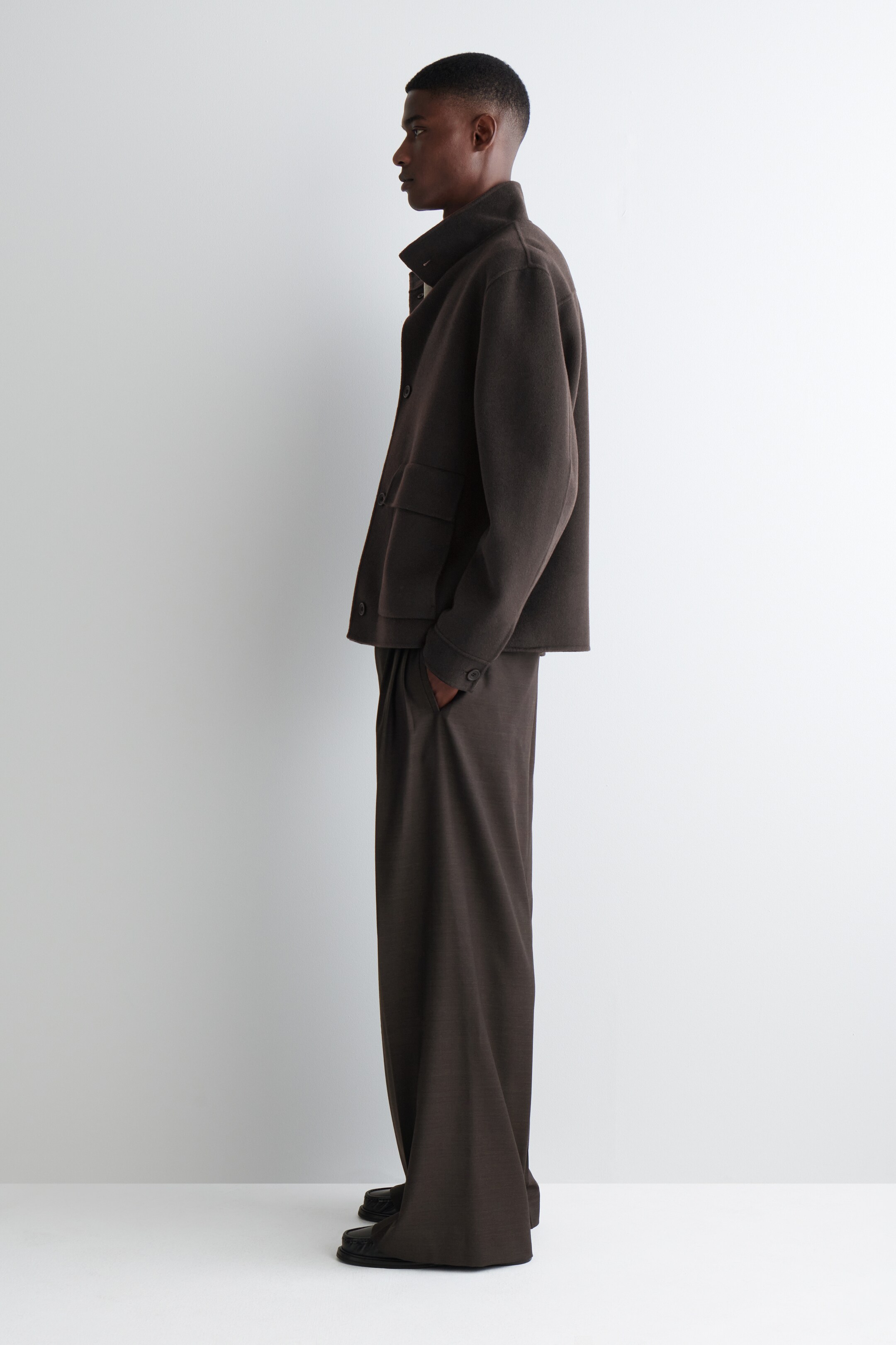Ver imagem maior: Um casaco Harrington castanho escuro está a ser usado por um modelo, que está de lado. O casaco apresenta uma gola alta em funil, um fecho de botões visível, um bolso lateral com aba e punhos com botões. O modelo também está a usar calças castanhas escuras a condizer e tem uma mão num bolso das calças.