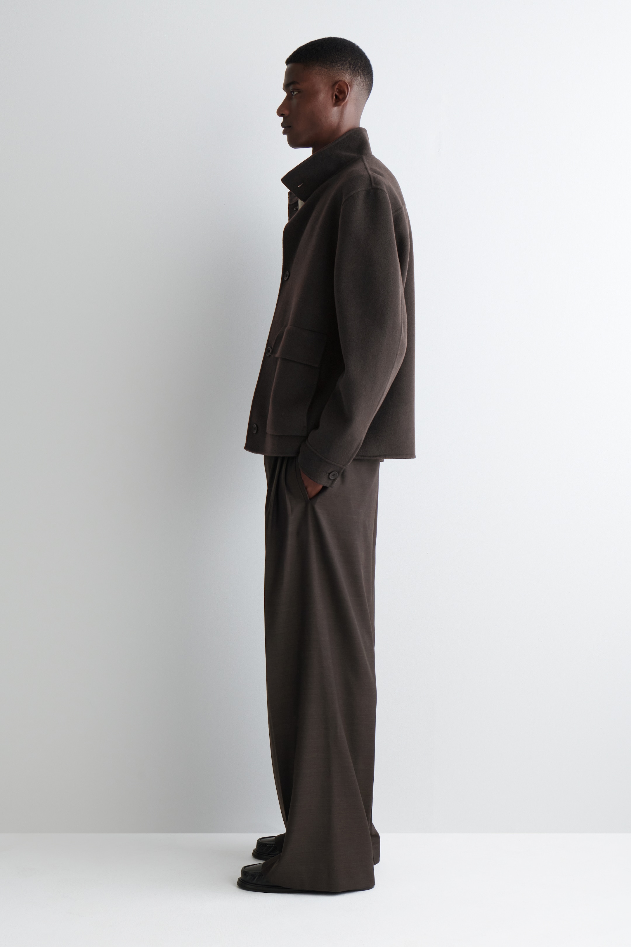 Grösseres Bild ansehen: Eine dunkelbraune Harrington-Jacke wird von einem Modell getragen, das seitlich steht. Die Jacke verfügt über einen hohen Trichterkragen, einen sichtbaren Knopfverschluss, eine seitliche Klappentasche und geknöpfte Manschetten. Das Modell trägt auch passende dunkelbraune Hosen und hat eine Hand in einer Hosentasche.