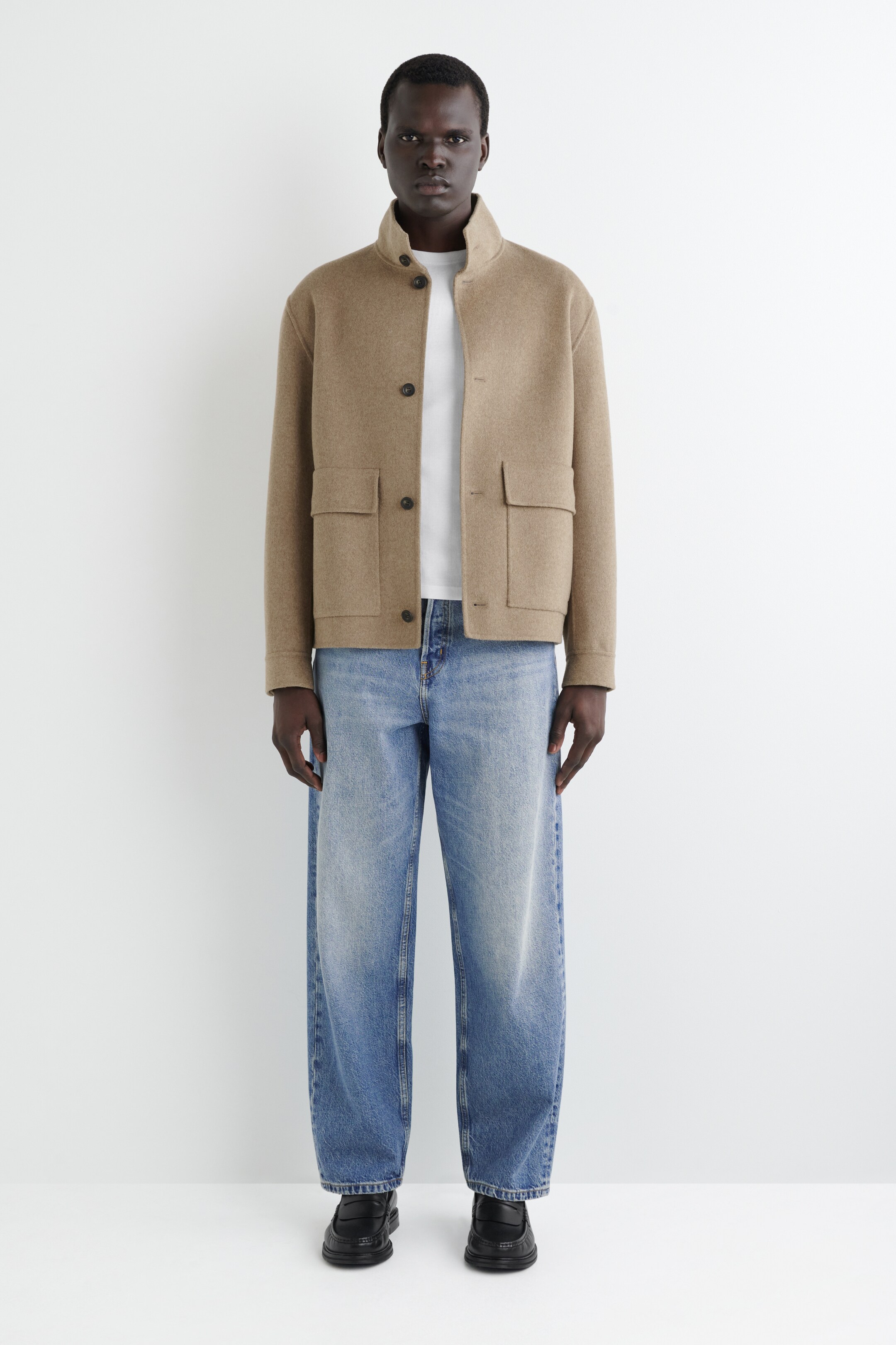Visualizza immagine più grande: Una giacca color tan con colletto a stand-up è presentata frontalmente, caratterizzata da una chiusura con bottoni sul davanti e due grandi tasche con patta sui fianchi. La giacca è indossata sopra una t-shirt bianca con collo a giro, che si abbina a jeans a gamba larga azzurri con bottoni a vista, e mocassini neri con suola spessa.