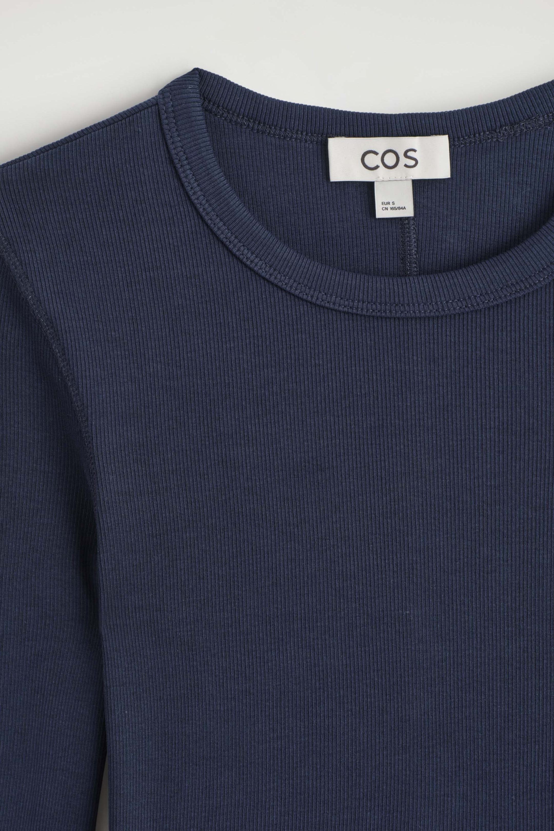 Showing a close-up of the navy blue slim ribbed cotton long-sleeved top from the front, highlighting the crew neckline and the fine vertical ribbed texture of the fabric. A white brand label is stitched inside the collar, displaying the brand name. Showing a close-up of the navy blue slim ribbed cotton long-sleeved top from the front, highlighting the crew neckline and the fine vertical ribbed texture of the fabric. A white brand label is stitched inside the collar, displaying the brand name.