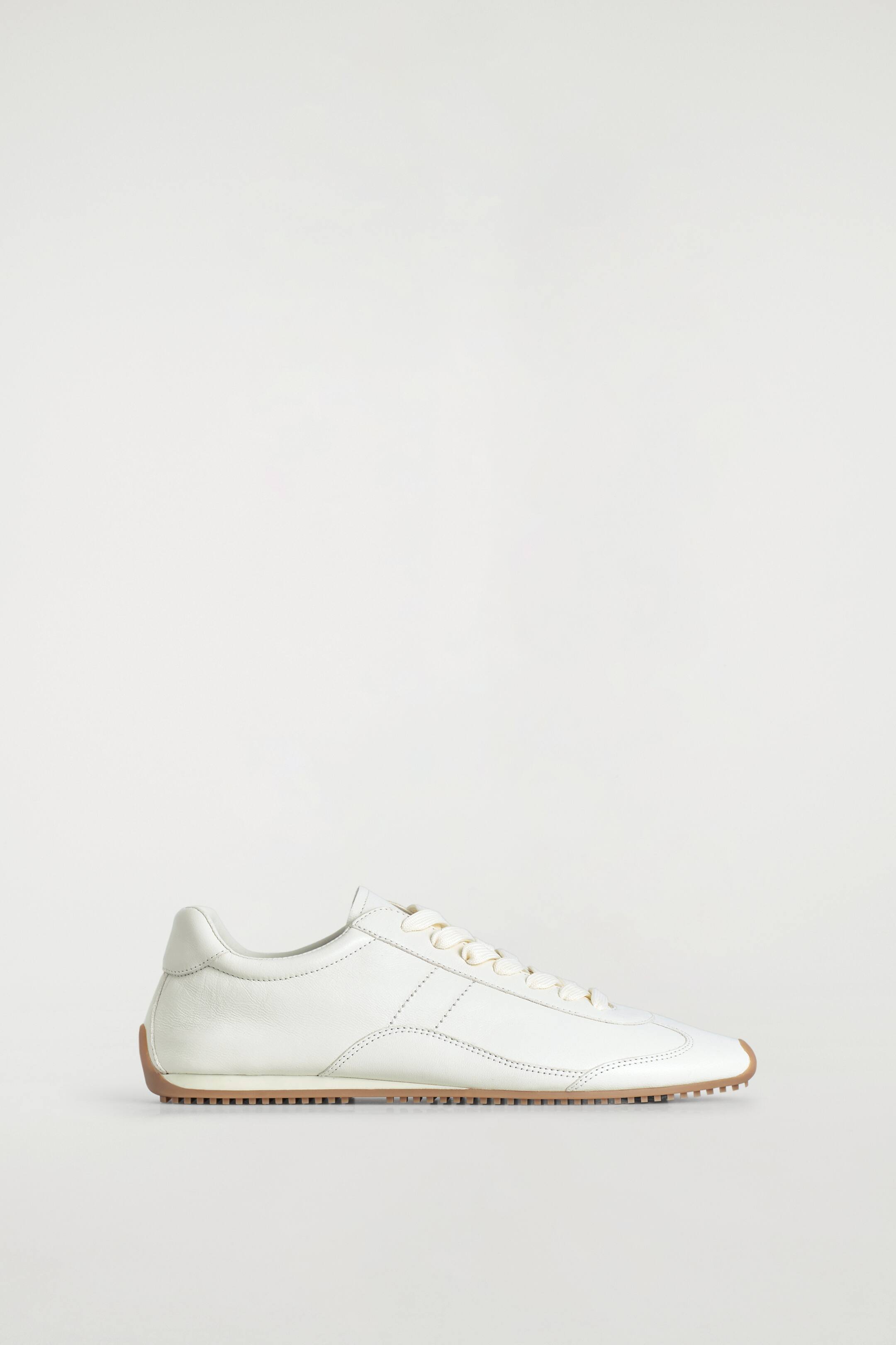 Agrandir l'image: Une sneaker en cuir blanc cassé est présentée de profil, avec une silhouette épurée, des surpiqûres ton sur ton, des lacets crème et une semelle en caoutchouc marron crantée contrastante.