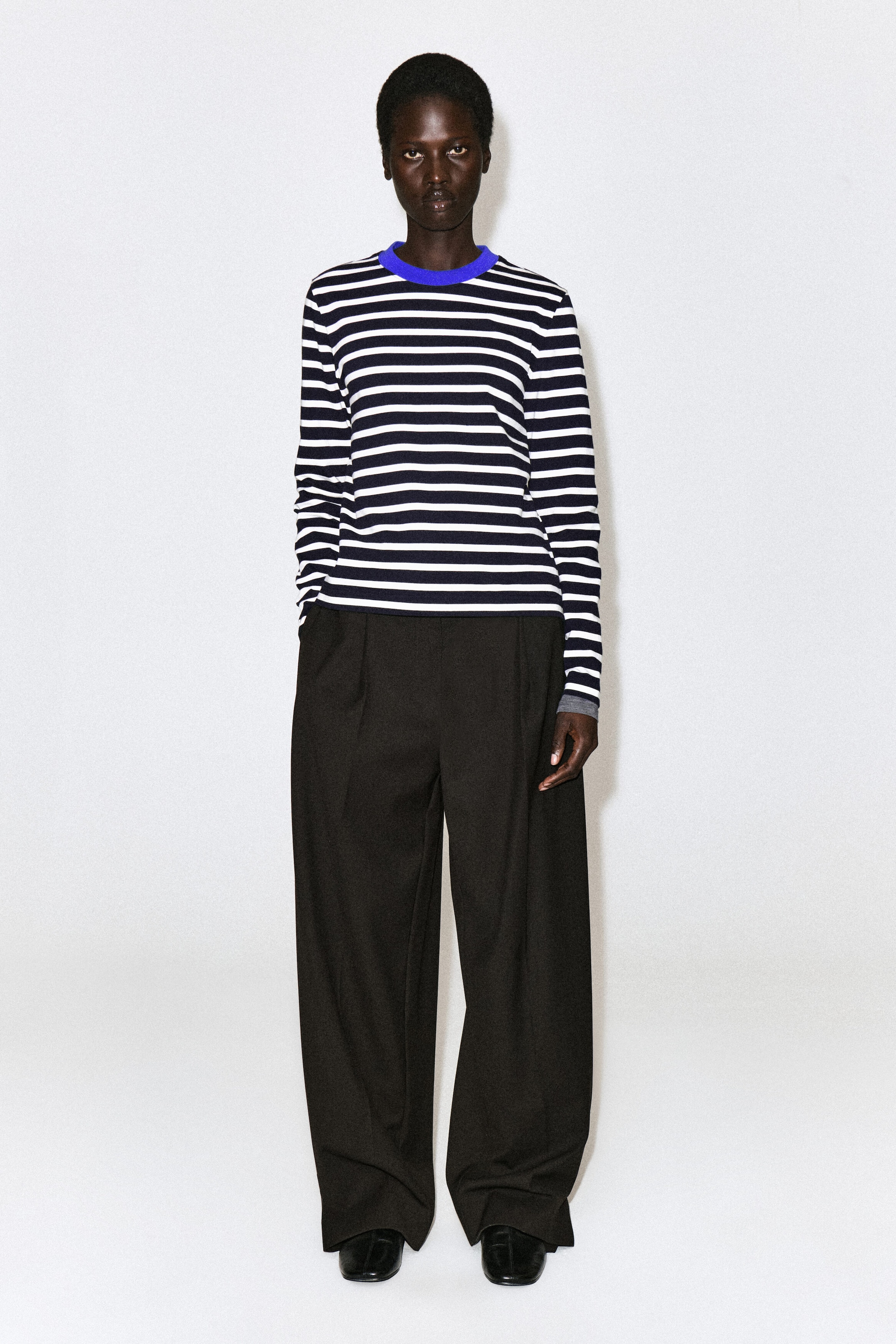 Ein schlicht geschnittenes Langarm-T-Shirt mit marineblauen und weißen horizontalen Streifen und einem kontrastierenden, leuchtend blauen Rundhalsausschnitt wird vom Model getragen. Das T-Shirt ist mit einer weiten schwarzen Hose kombiniert. Ein schlicht geschnittenes Langarm-T-Shirt mit marineblauen und weißen horizontalen Streifen und einem kontrastierenden, leuchtend blauen Rundhalsausschnitt wird vom Model getragen. Das T-Shirt ist mit einer weiten schwarzen Hose kombiniert.