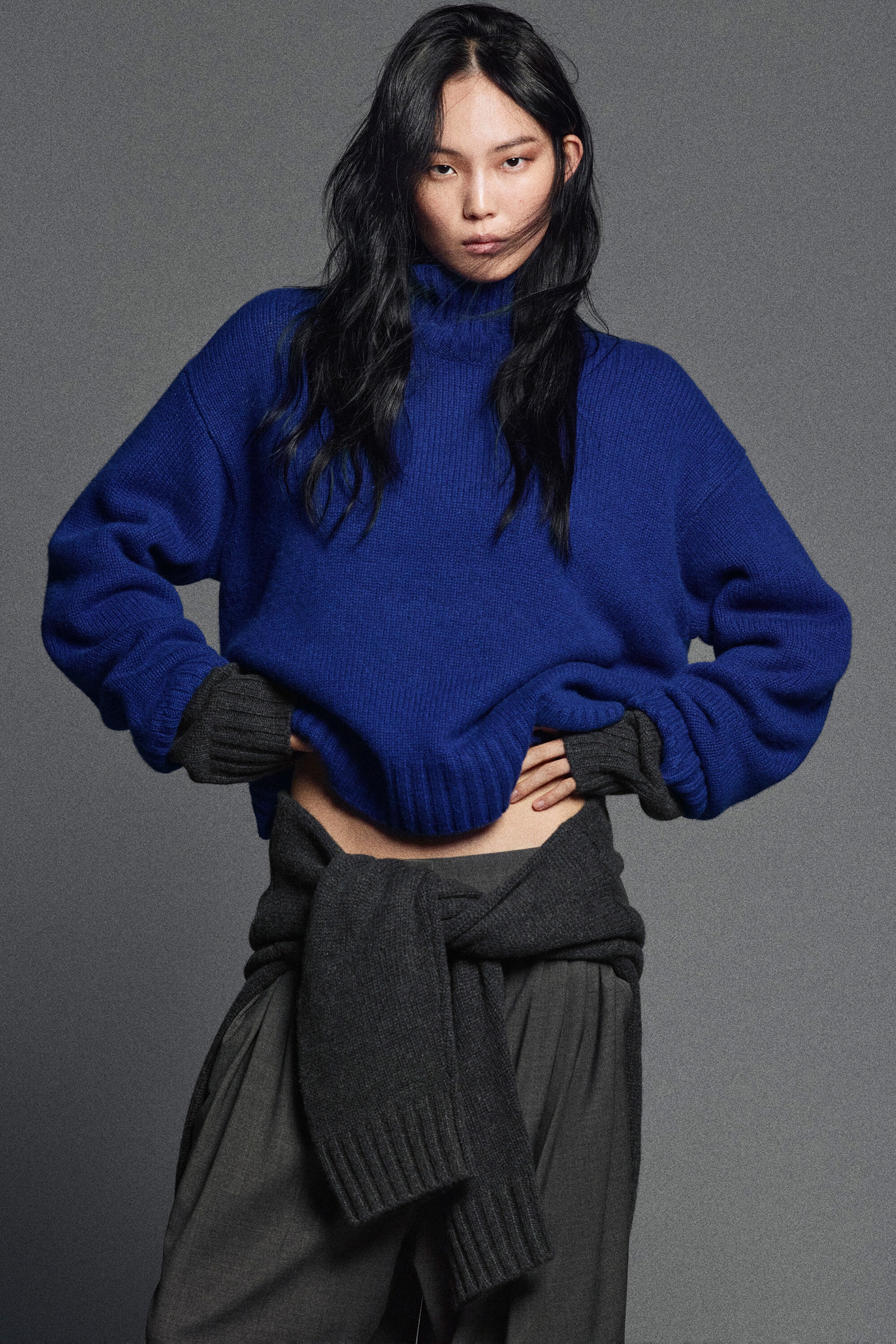 Shown from the front, a model is wearing a vibrant blue, chunky cashmere funnel-neck jumper with a relaxed fit, paired with dark gray pleated mélange-wool trousers. A dark gray knit sweater is tied around the waist, and matching dark gray ribbed cuffs are peeking out from the jumper's sleeves.