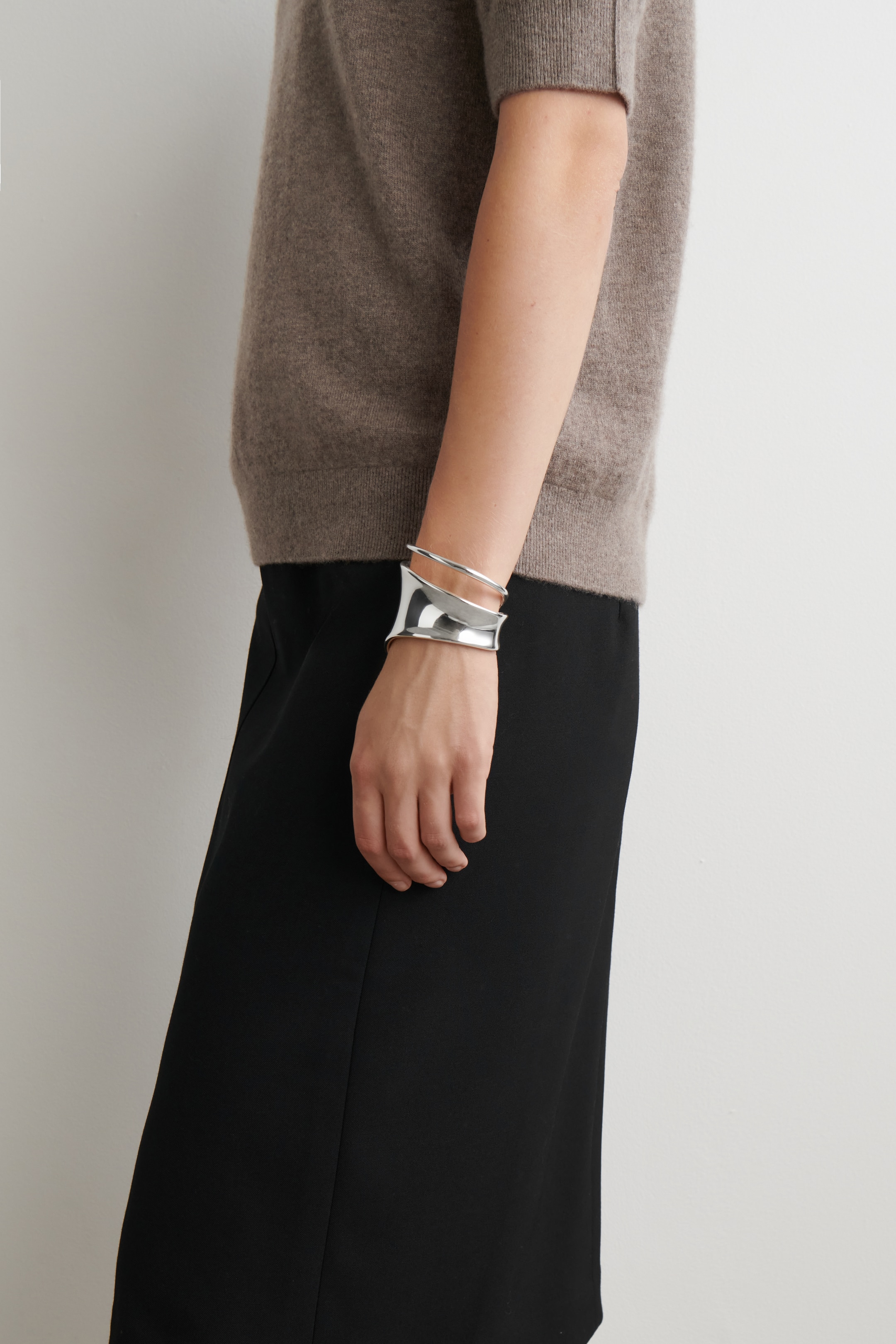 View larger image: A wide, polished silver cuff bracelet is being worn on a wrist, presented from a side angle. Its sculptural design is displaying undulating, overlapping forms that are wrapping around the arm.
