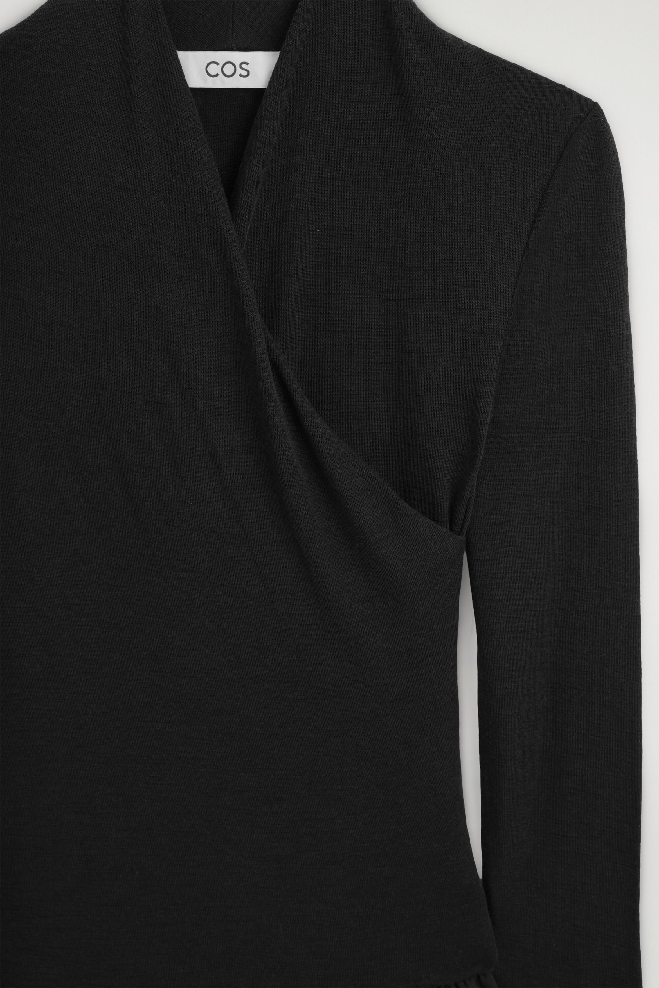 The black contrast-panel wrap-over dress is shown in a close-up front view, featuring a draped wrap-over design that creates a V-neckline. The garment displays a fine-knit texture and long sleeves, with a brand label visible at the inner collar. The black contrast-panel wrap-over dress is shown in a close-up front view, featuring a draped wrap-over design that creates a V-neckline. The garment displays a fine-knit texture and long sleeves, with a brand label visible at the inner collar.