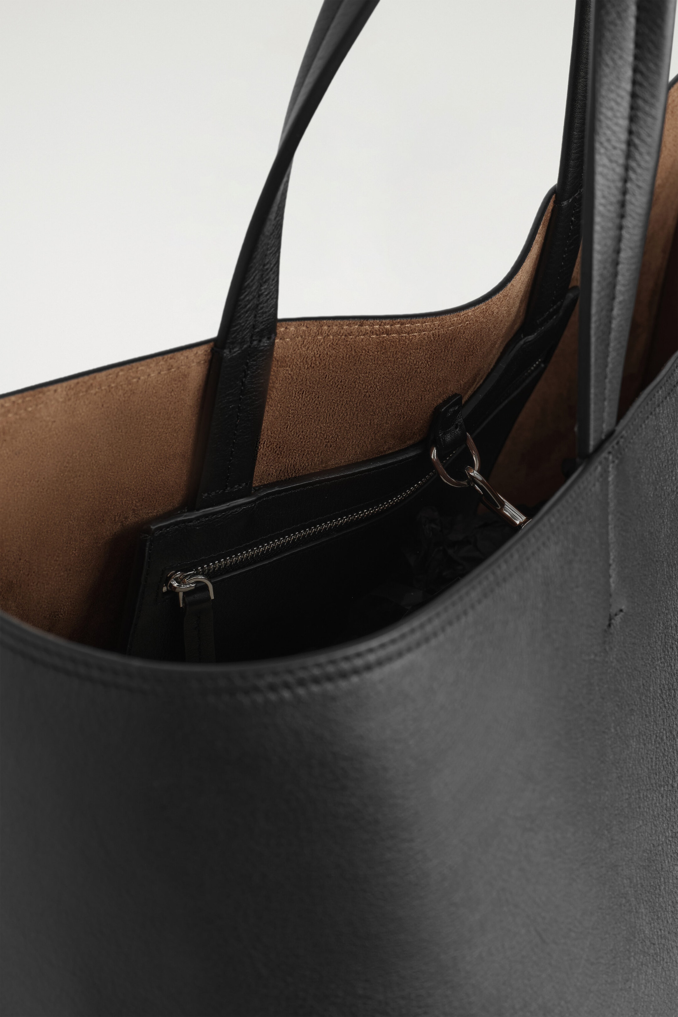 A black leather tote bag is shown from above, revealing its interior with a textured black exterior and a contrasting tan suede lining. A matching black leather zippered pouch hangs inside, attached to the interior wall with a silver-tone metal clasp. A black leather tote bag is shown from above, revealing its interior with a textured black exterior and a contrasting tan suede lining. A matching black leather zippered pouch hangs inside, attached to the interior wall with a silver-tone metal clasp.