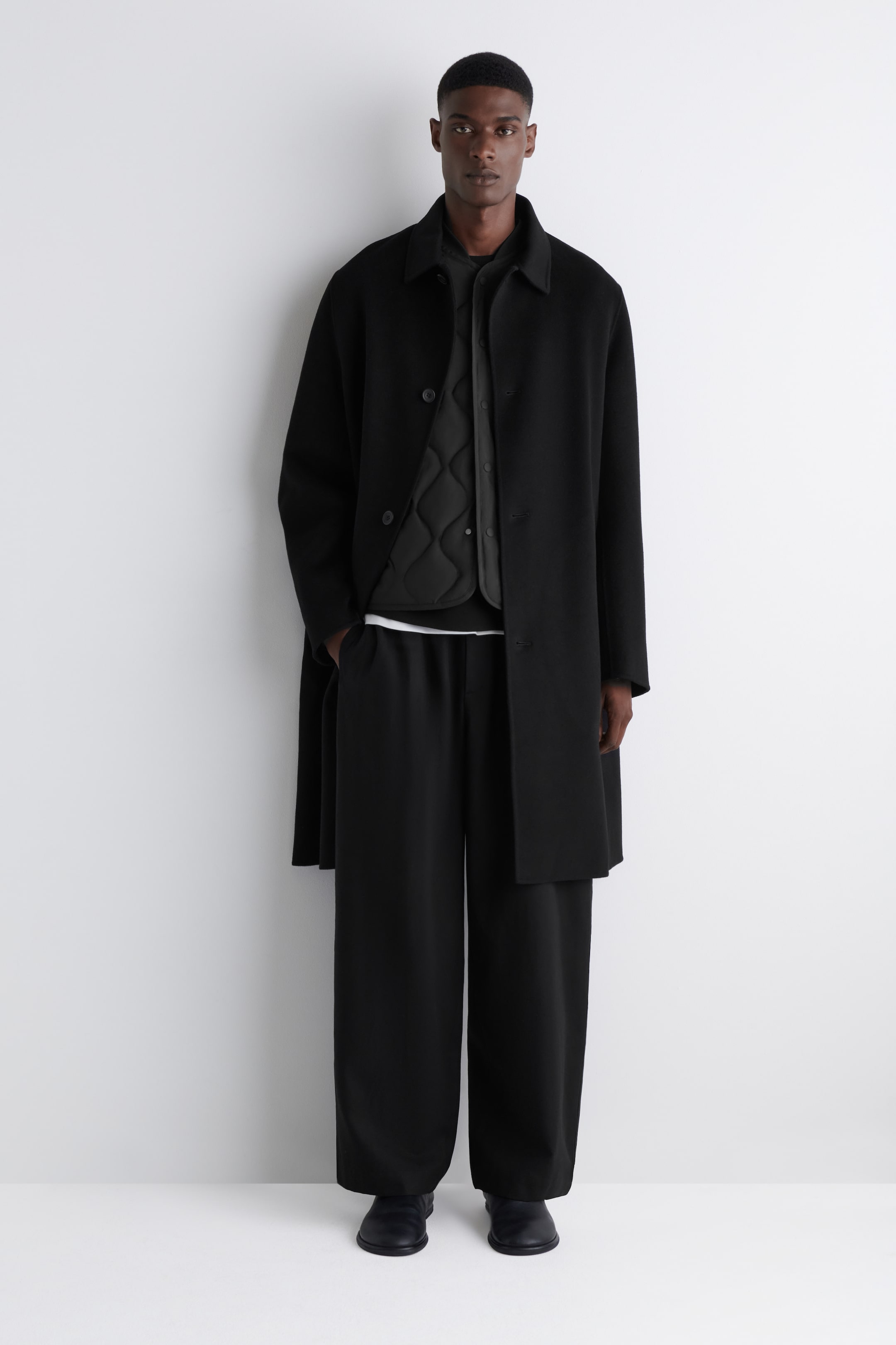 Größeres Bild anzeigen: Ein komplett schwarzes Outfit wird an einer Person präsentiert, die nach vorne schaut. Ein langer, schwarzer Mantel mit Kragen, der mit drei sichtbaren Knöpfen geschlossen ist, wird über einer schwarzen, gesteppten Weste getragen. Ein schwarzes Oberteil, das einen weißen Saum darunter zeigt, ist unter der Weste geschichtet und ergänzt schwarze, weit geschnittene Hosen und schwarze minimalistische Schuhe.