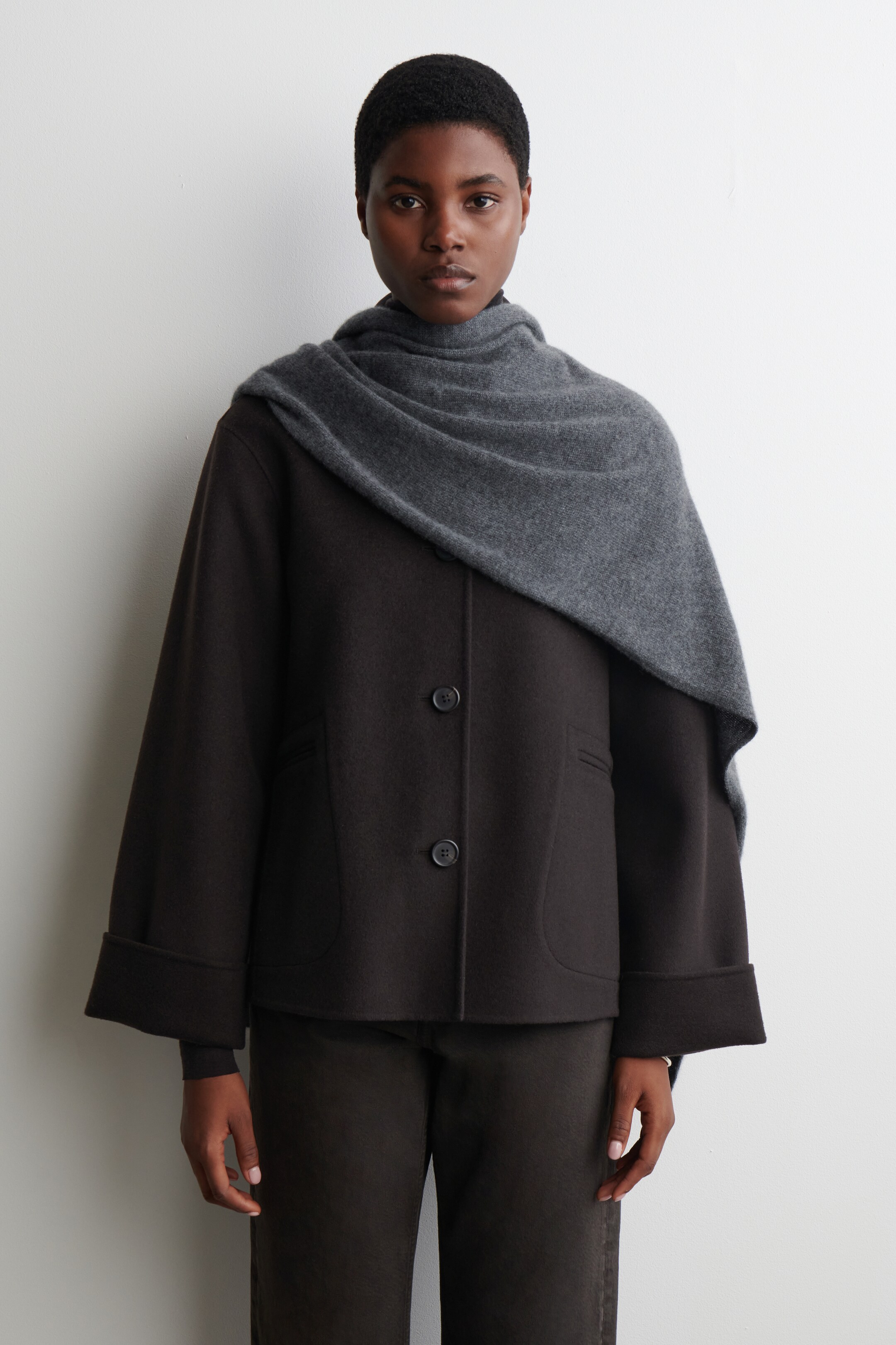 View larger image: A dark brown jacket is being presented on a person facing forward, featuring a two-button front closure and two rounded patch pockets. Its wide sleeves are being worn with large, rolled cuffs, and a gray scarf is being draped over its shoulders.