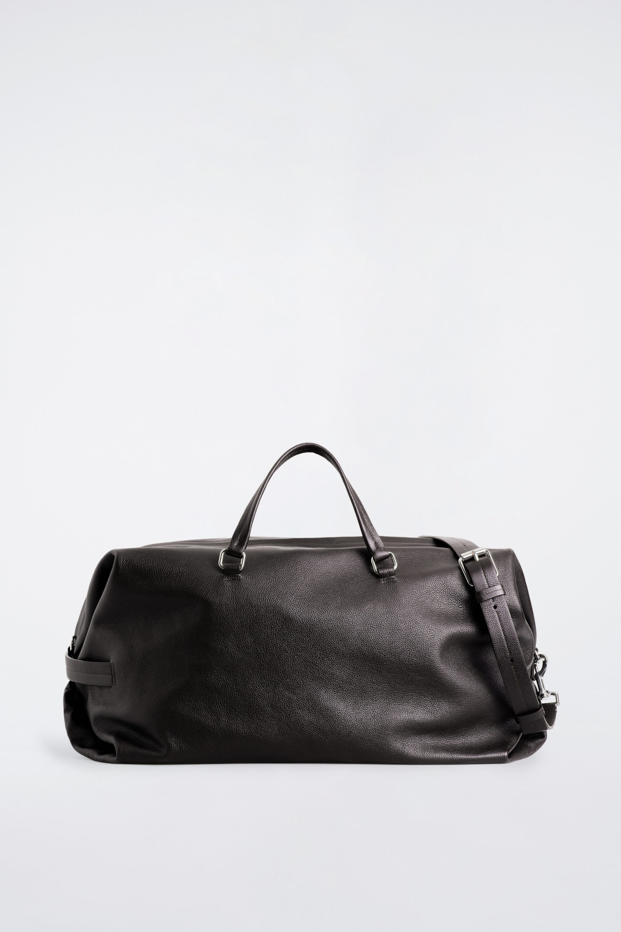 The Departure Duffle Bag in textured dark leather is shown in a front-facing view, showcasing its slouchy structure with dual rounded top handles attached by silver-toned metal hardware. An adjustable leather shoulder strap with a silver buckle drapes over the right-hand side of the bag.